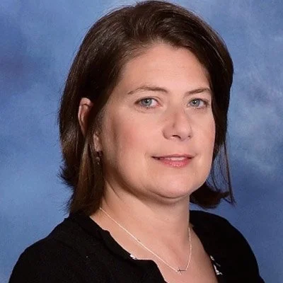 A woman with shoulder-length dark hair and light eyes, wearing a black top and a silver necklace, posing against a blue background.