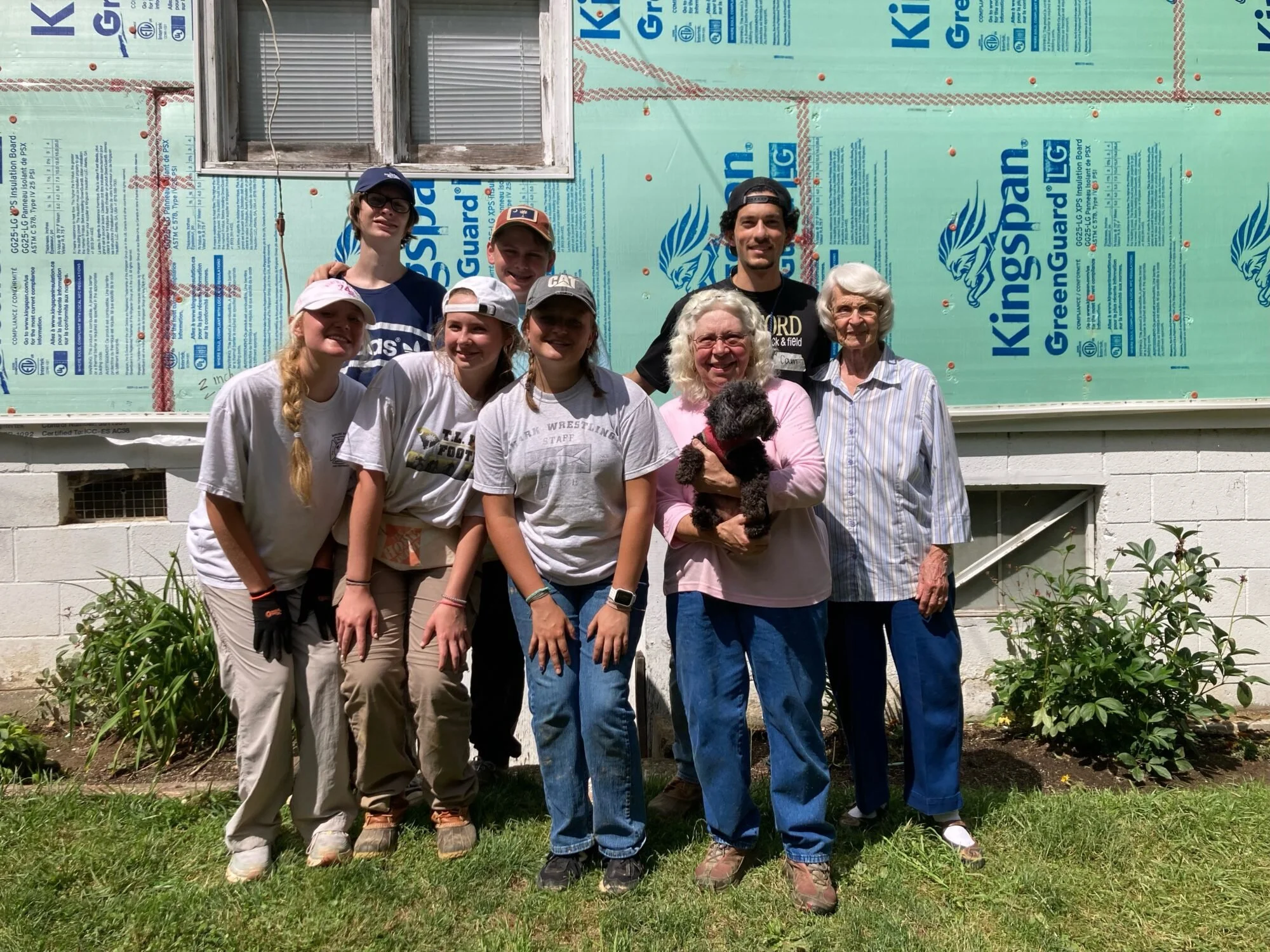 Group of nine people standing outside in front of a house under construction, with insulation visible on the wall. They are smiling and some are holding a small black dog. The group includes older and younger women and men, dressed casually in t-shirts and jeans or khakis.