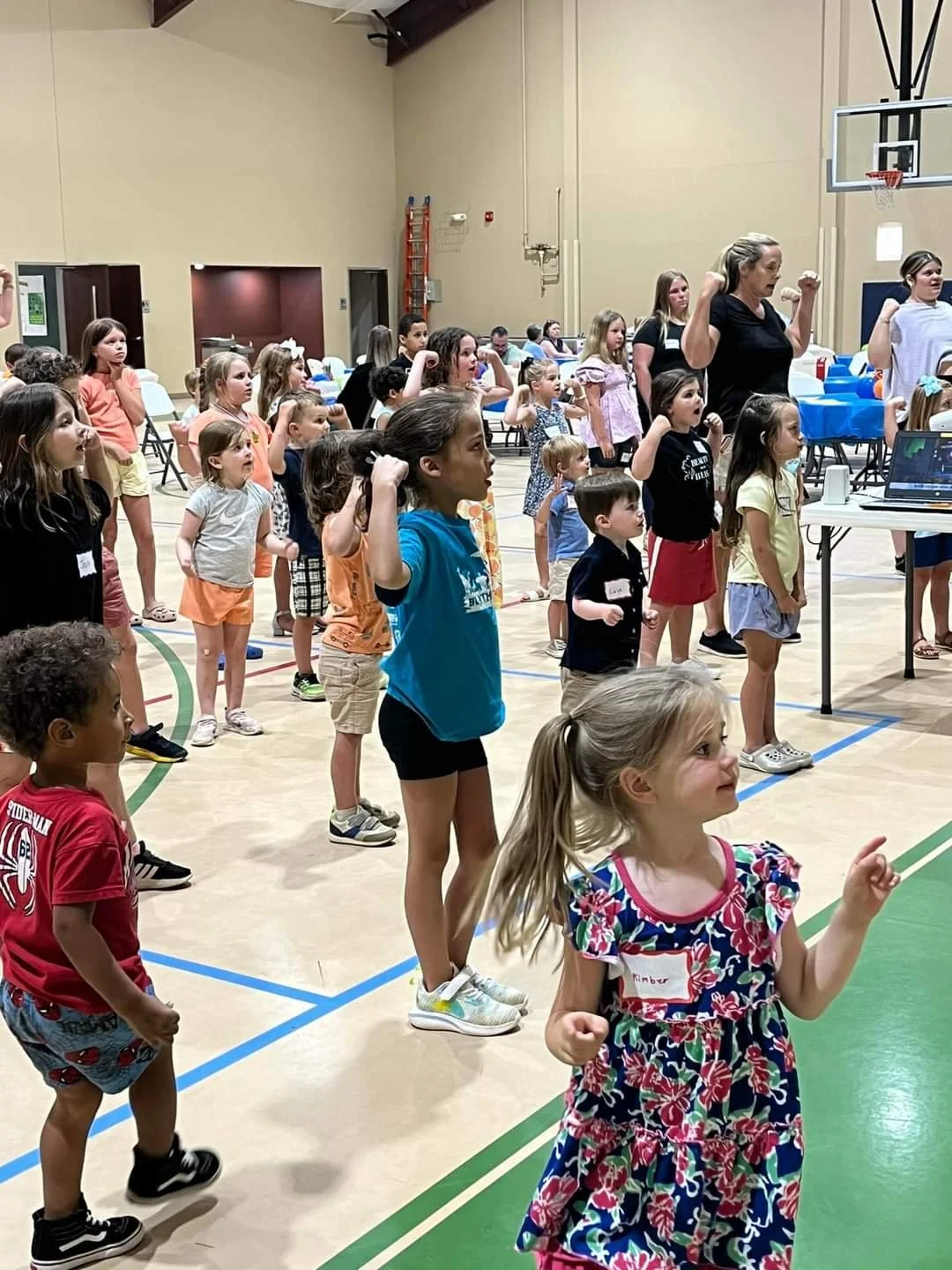 Children and instructors participating in a group activity or fitness class inside a gymnasium, with kids standing on a gym floor and raising their fists in the air, led by an instructor.