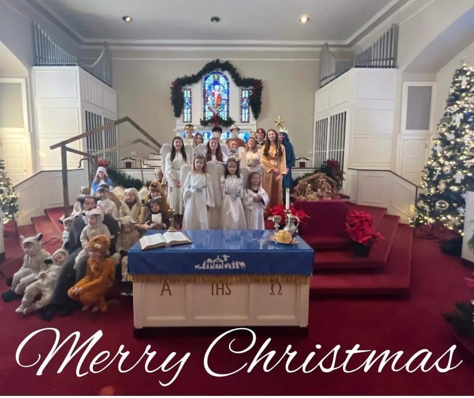 Children and adults dressed as angels, animals, and other characters in a Christmas pageant inside a church, with decorated Christmas trees, lights, and a stained glass window in the background. The bottom of the image has the greeting "Merry Christmas."