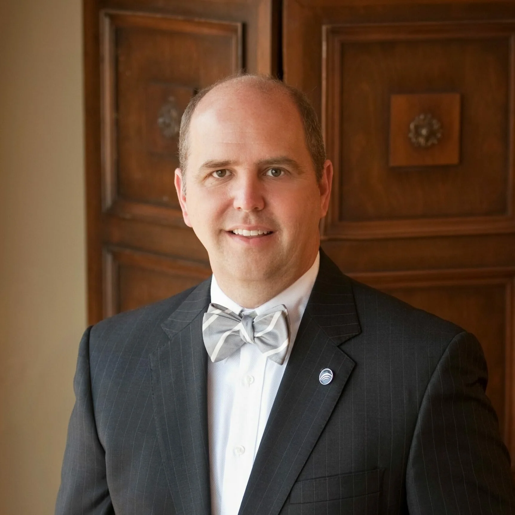 Portrait of a man in a dark suit with a bow tie, standing indoors in front of a wooden panel wall.