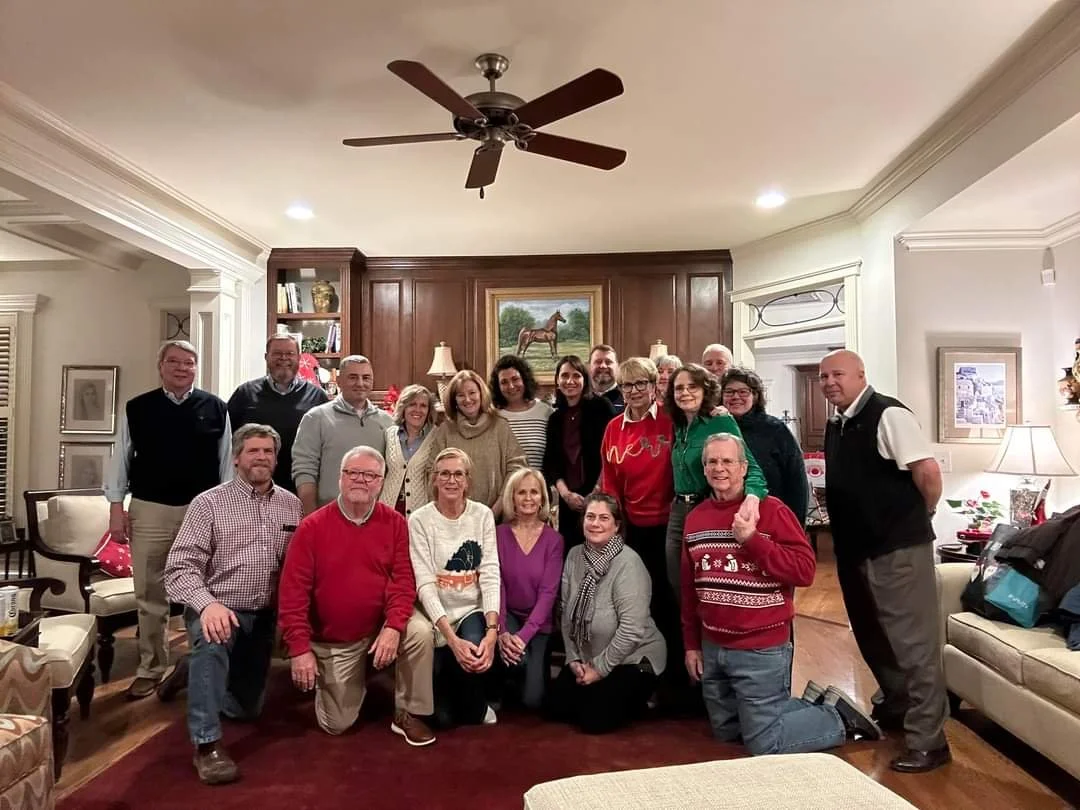 Group of 16 people gathered in a living room, posing for a photo. The room has a fireplace with decorative items and a painting of a horse hung above it. The room is decorated for the holiday season.