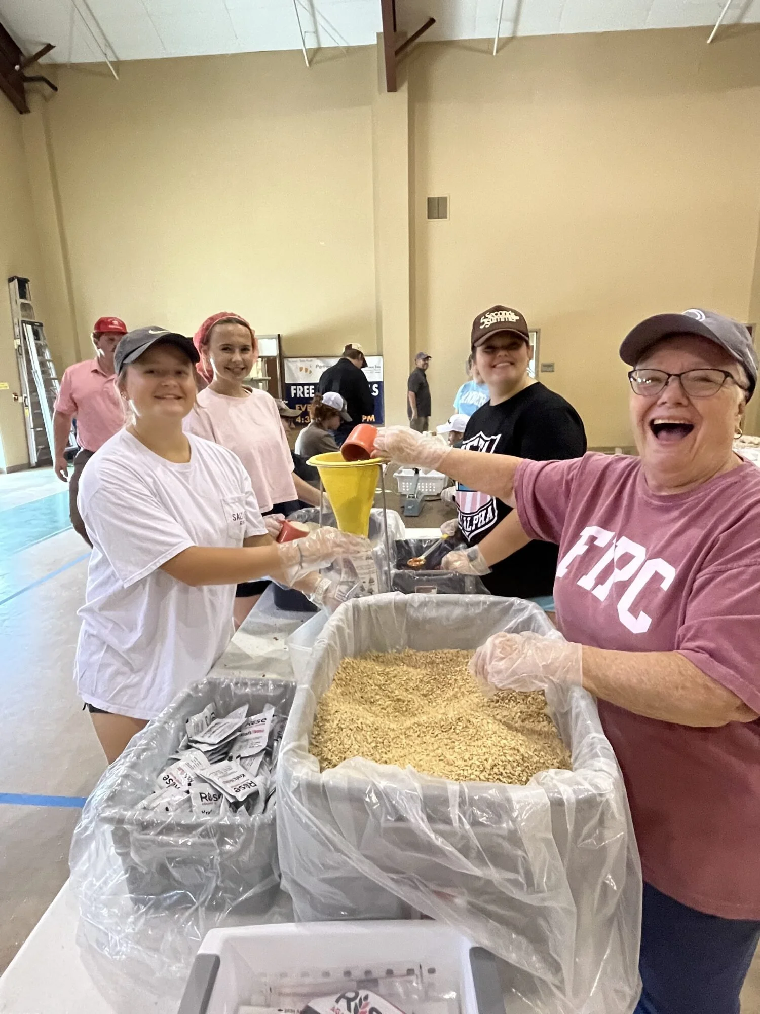 Group of people serving food at a community event in a large indoor space, with tables, large trash containers, and volunteers wearing gloves.