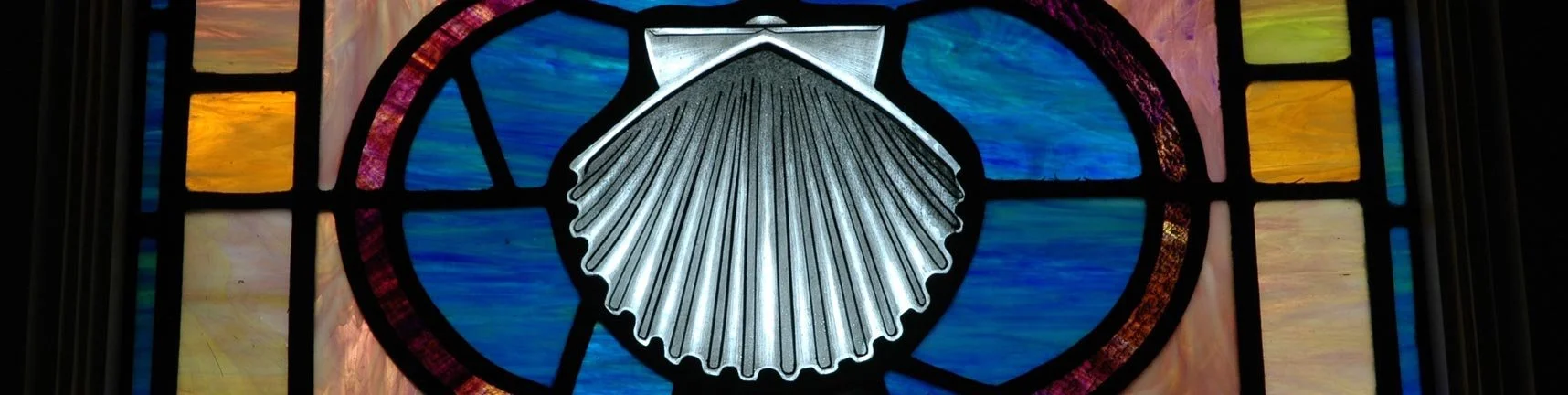 A stained glass window featuring a scallop shell design with colorful blue, yellow, purple, and orange sections.