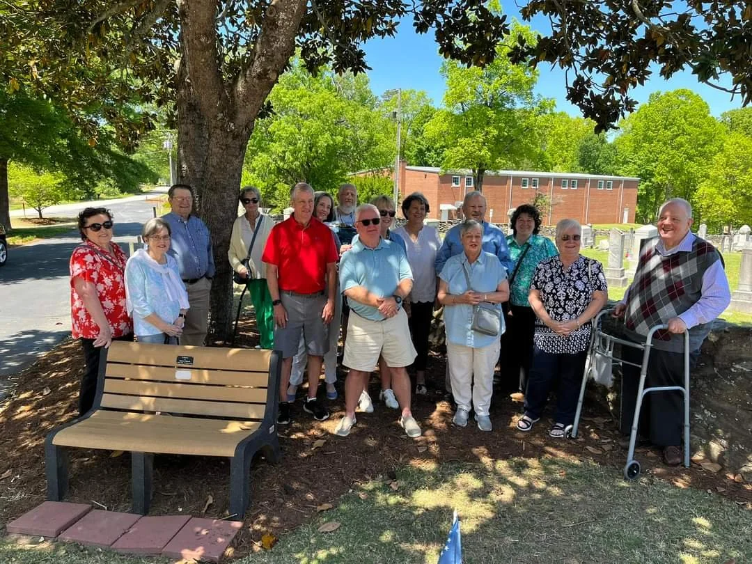 A group of fourteen people, mostly seniors, standing under a large tree in a park or cemetery on a sunny day. Some are smiling and wearing sunglasses, with a few using walkers. Background includes green trees, a brick building, and grave markers.