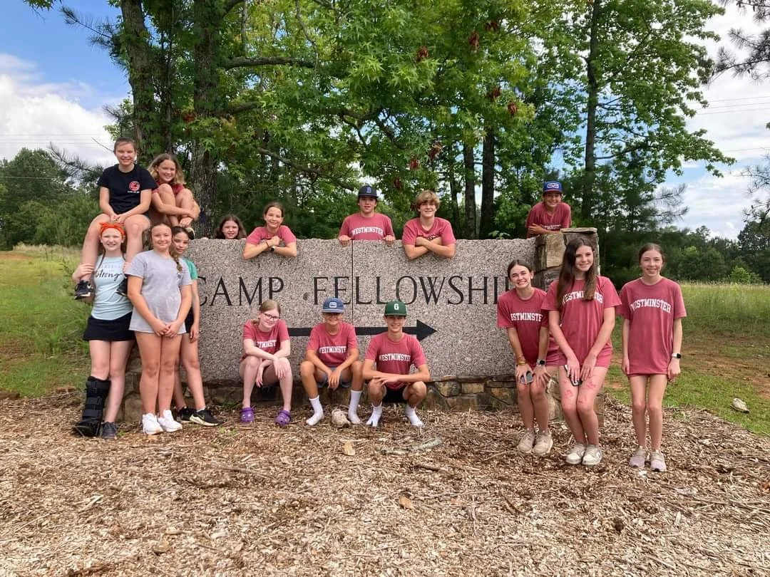 Group of children at Camp Fellowship, some sitting and some standing in front of a large stone sign with the words 'CAMP FELLOWSHIP' engraved. They are outdoors with trees and a cloudy sky in the background.