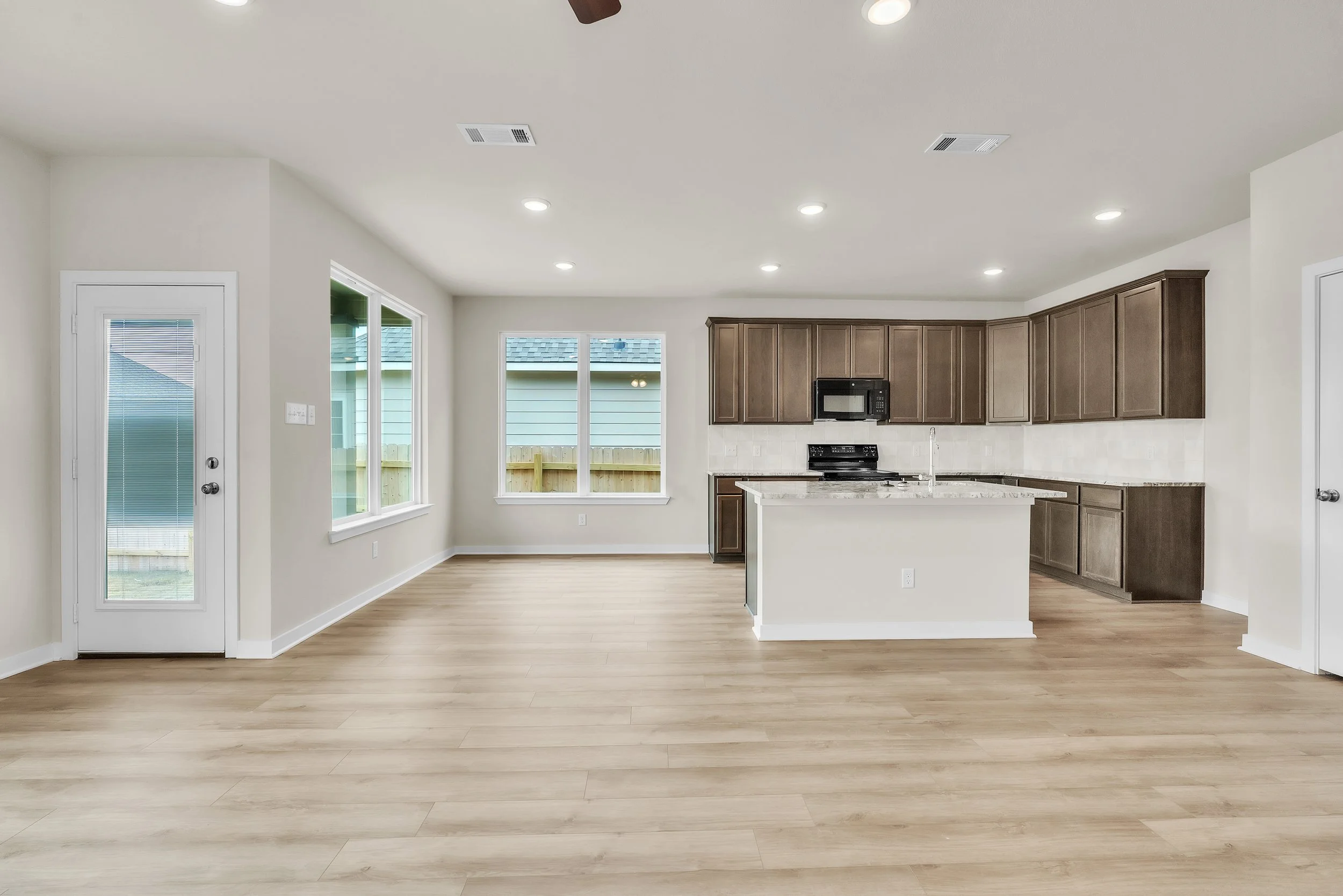 Empty modern kitchen with light wood floors, beige walls, dark wood cabinets, granite island, stainless steel appliances, and large windows with a view of a neighboring house and backyard.