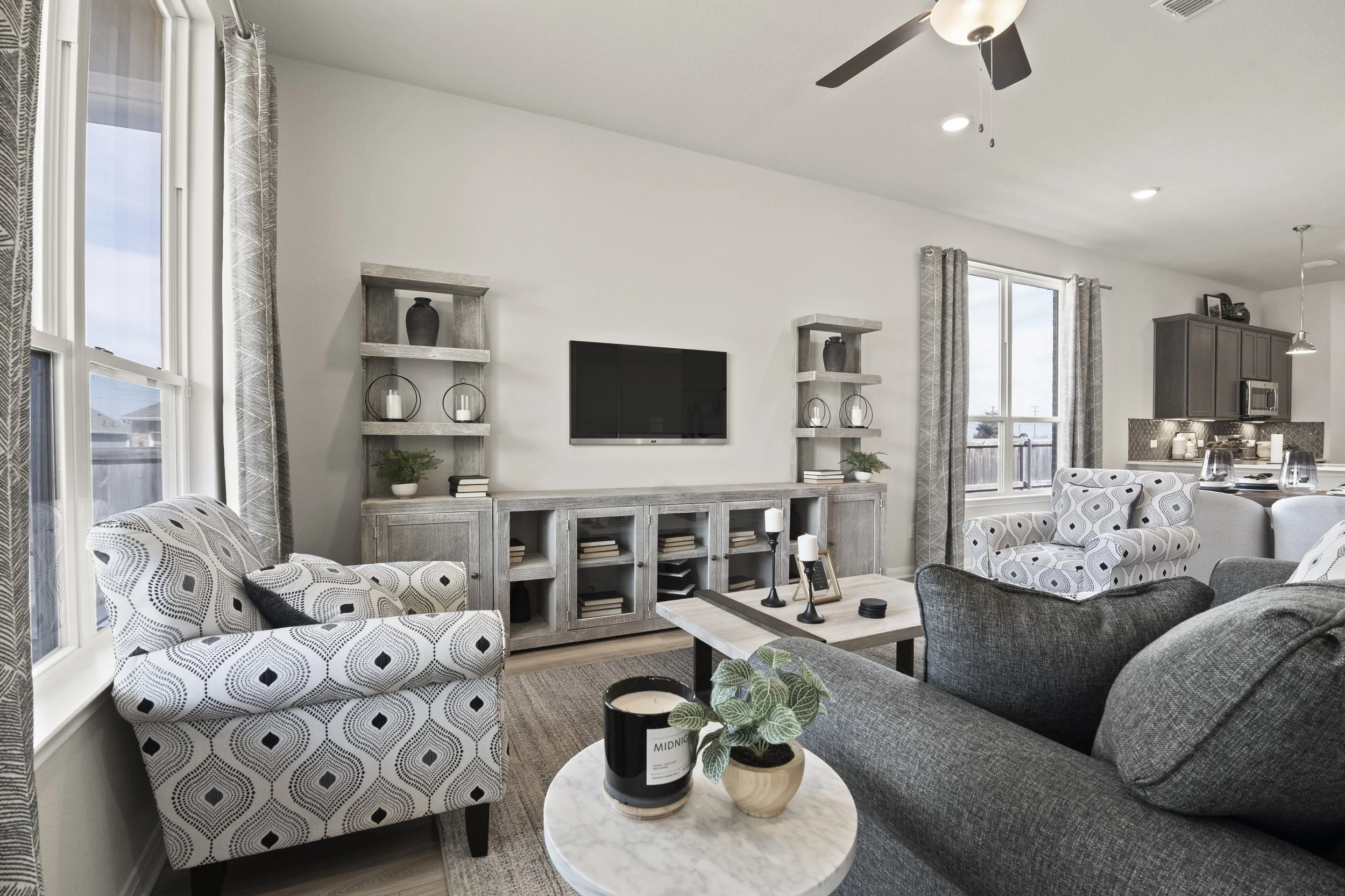 Living room with a gray sofa, patterned armchairs, a wooden coffee table, a TV on the wall, and large windows with curtains. Part of the kitchen is visible in the background.