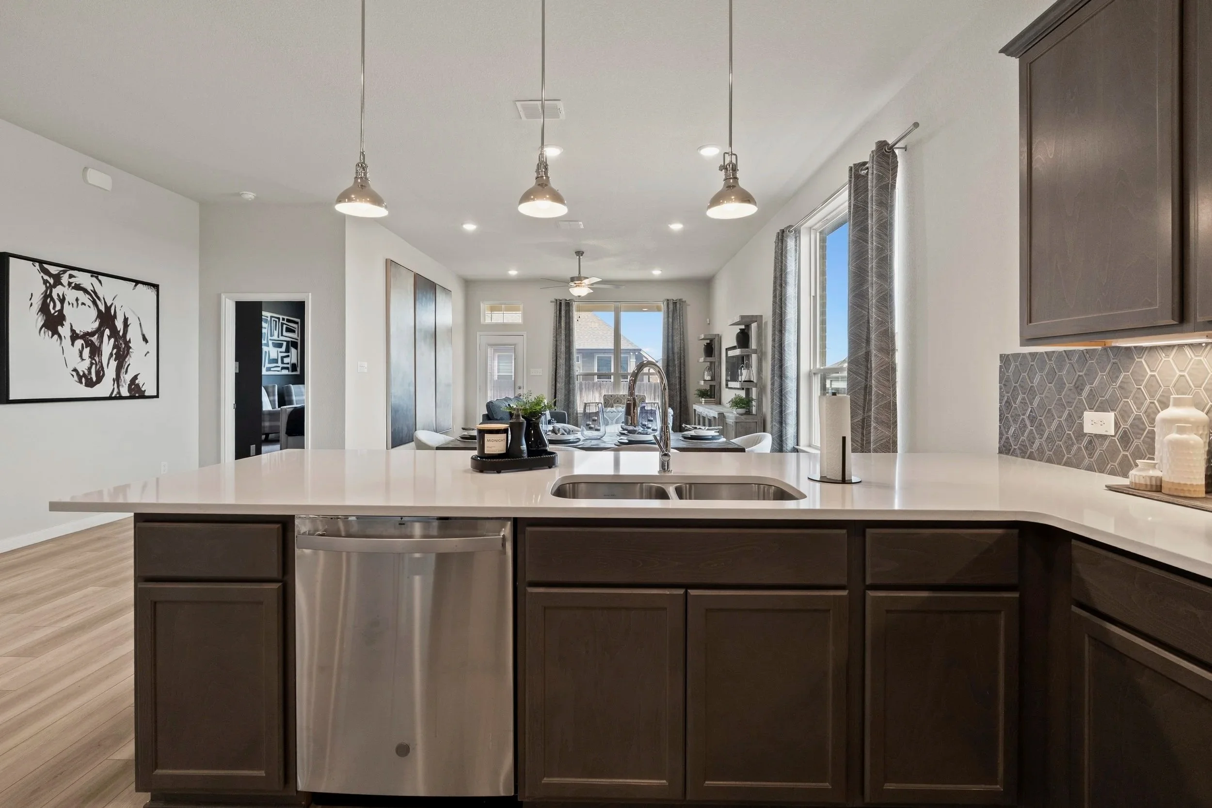 Open kitchen with dark cabinets, white countertop, stainless steel dishwasher, and a sink with a modern faucet. The background shows a dining area with a table set for a meal, large windows with curtains, and a living area with a ceiling fan and artw
