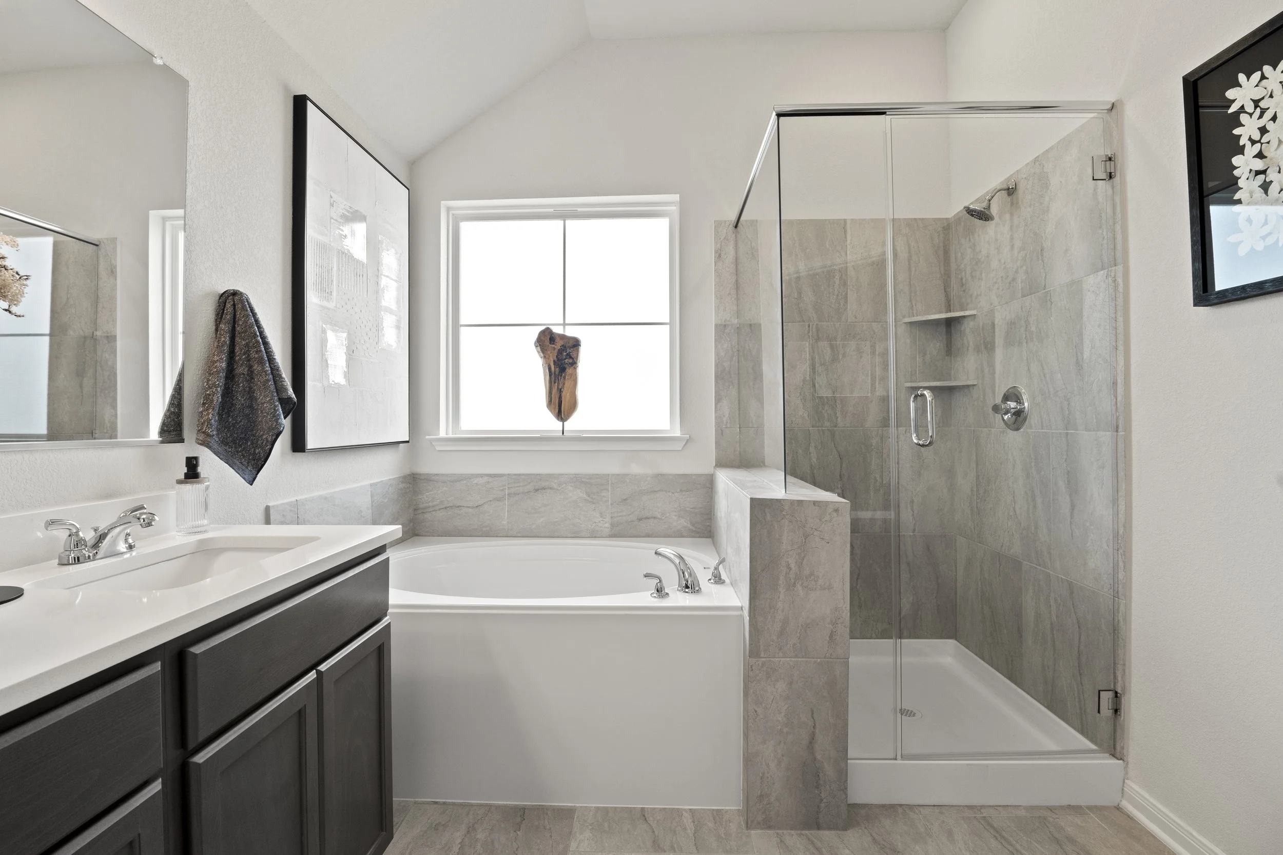 Modern bathroom with a white bathtub, a glass-enclosed shower, a dark vanity with a white countertop, a large mirror, a window with a decorative piece hanging, and framed artwork on the walls.