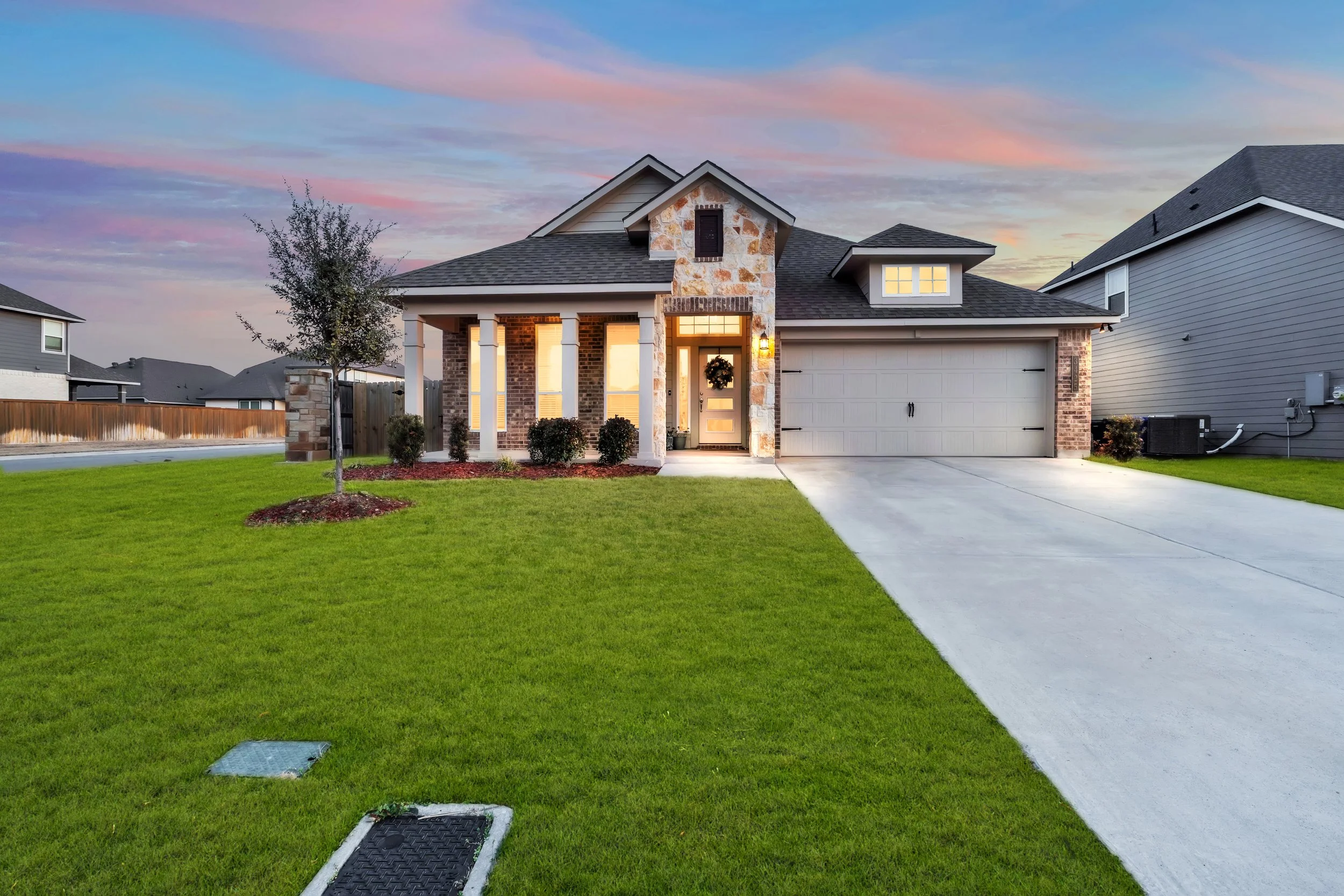 A modern house with a brick and stone facade, porch with four columns, and a double garage, illuminated during sunset with a landscaped lawn and a small tree in front.