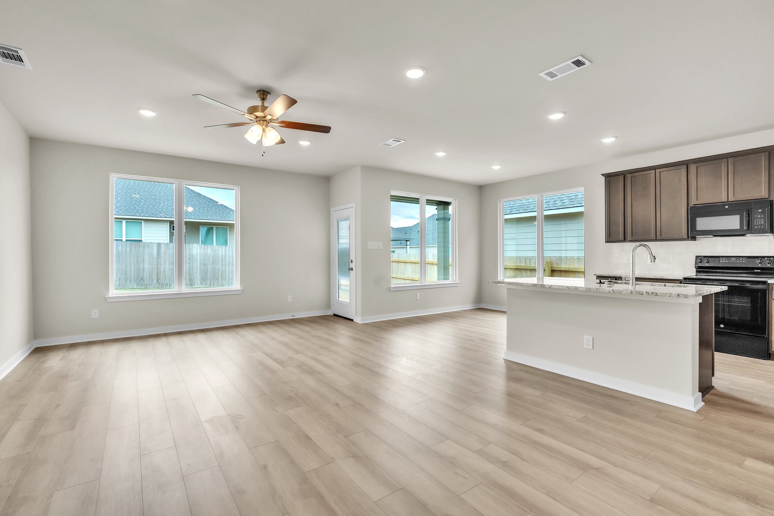 Empty living room and kitchen area in a modern home with light-colored flooring, white walls, a ceiling fan, large windows, and a kitchen with dark cabinets and black appliances.