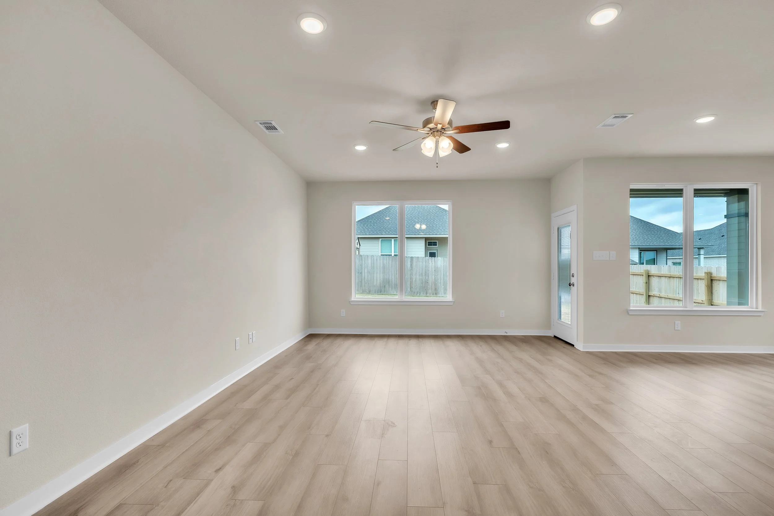 Empty living room with light wood flooring, white walls, multiple ceiling lights, a ceiling fan, and large windows showing a fenced backyard with neighboring houses.