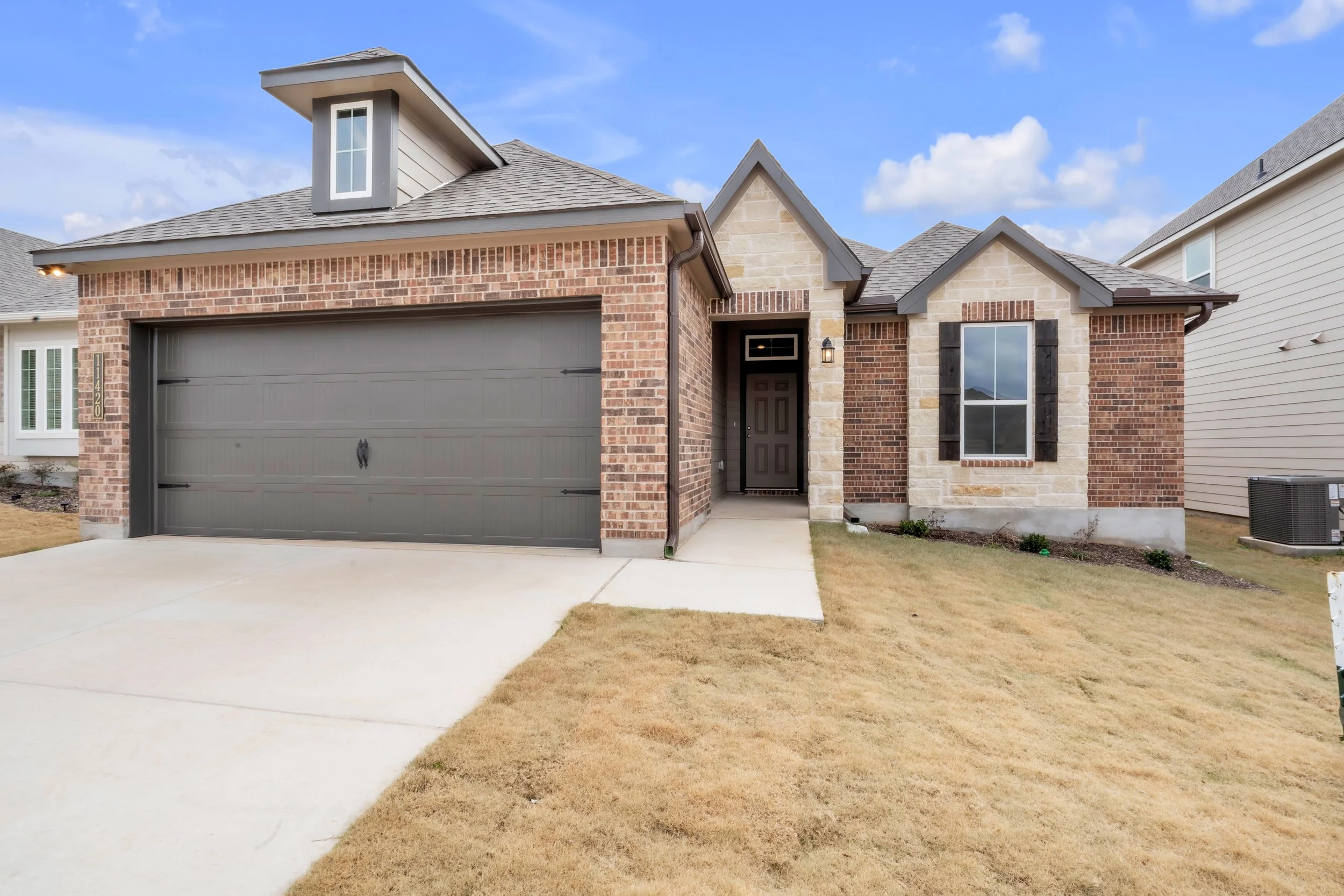 Front view of a modern suburban house with brick and stone exterior, gray garage door, black shutters, and a small front yard with brown grass.