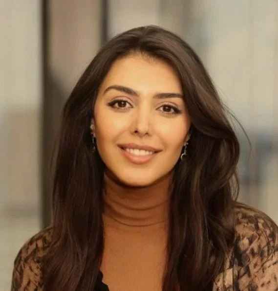 A woman with long dark hair, wearing a tan turtleneck and lace-patterned blazer, smiling indoors.
