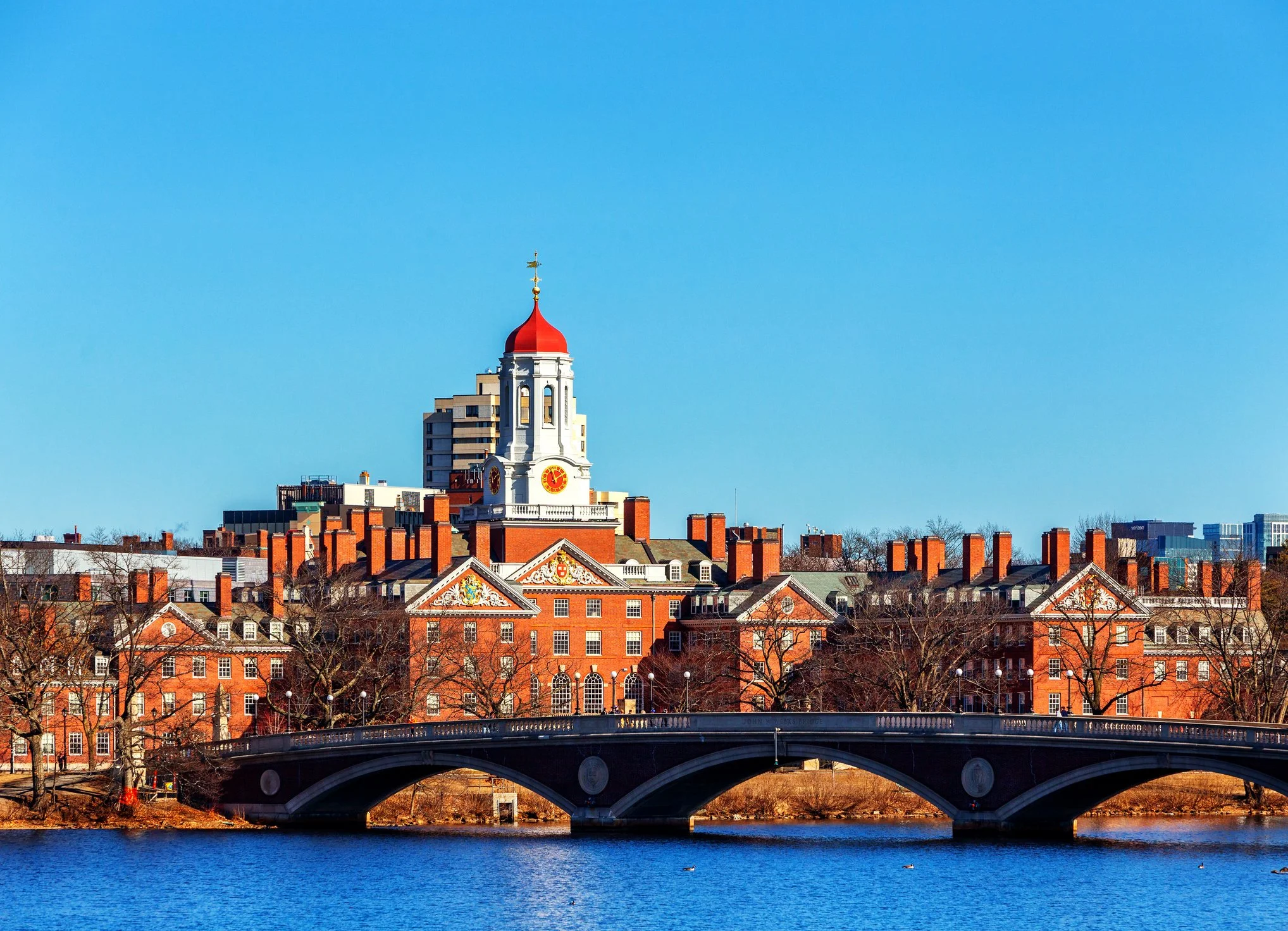 Historic brick buildings with a central white clock tower topped with a red dome and weather vane, across a river with a stone bridge and leafless trees against a clear blue sky.
