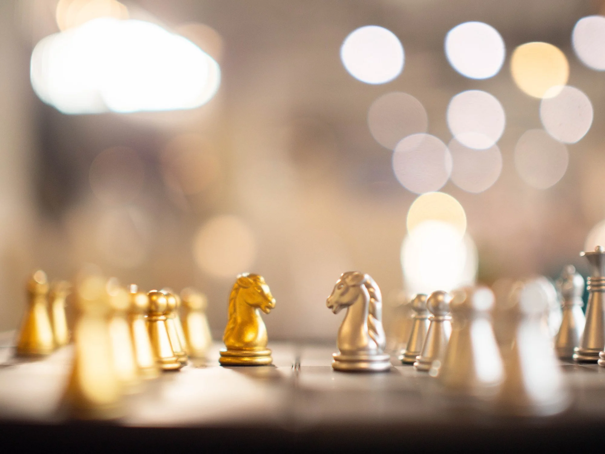 Close-up of gold and silver chess pieces on a chessboard, with a blurred background of bright city lights.