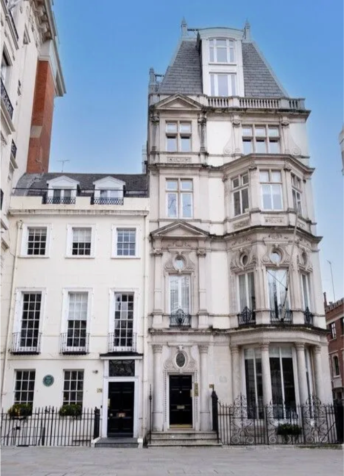 A tall, ornate, historic white building with multiple architectural details, including bay windows, decorative moldings, and a mansard roof, in an urban setting with neighboring buildings.