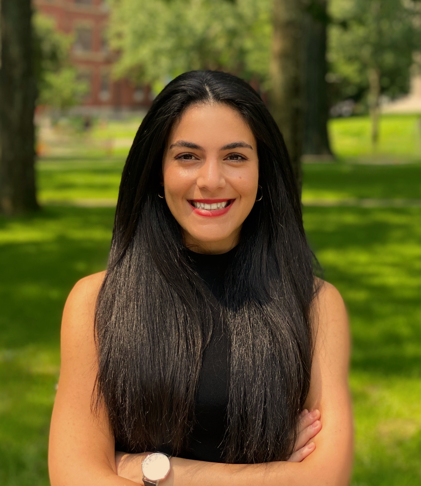 A woman with long dark hair smiling outdoors in a park with green trees and grass.