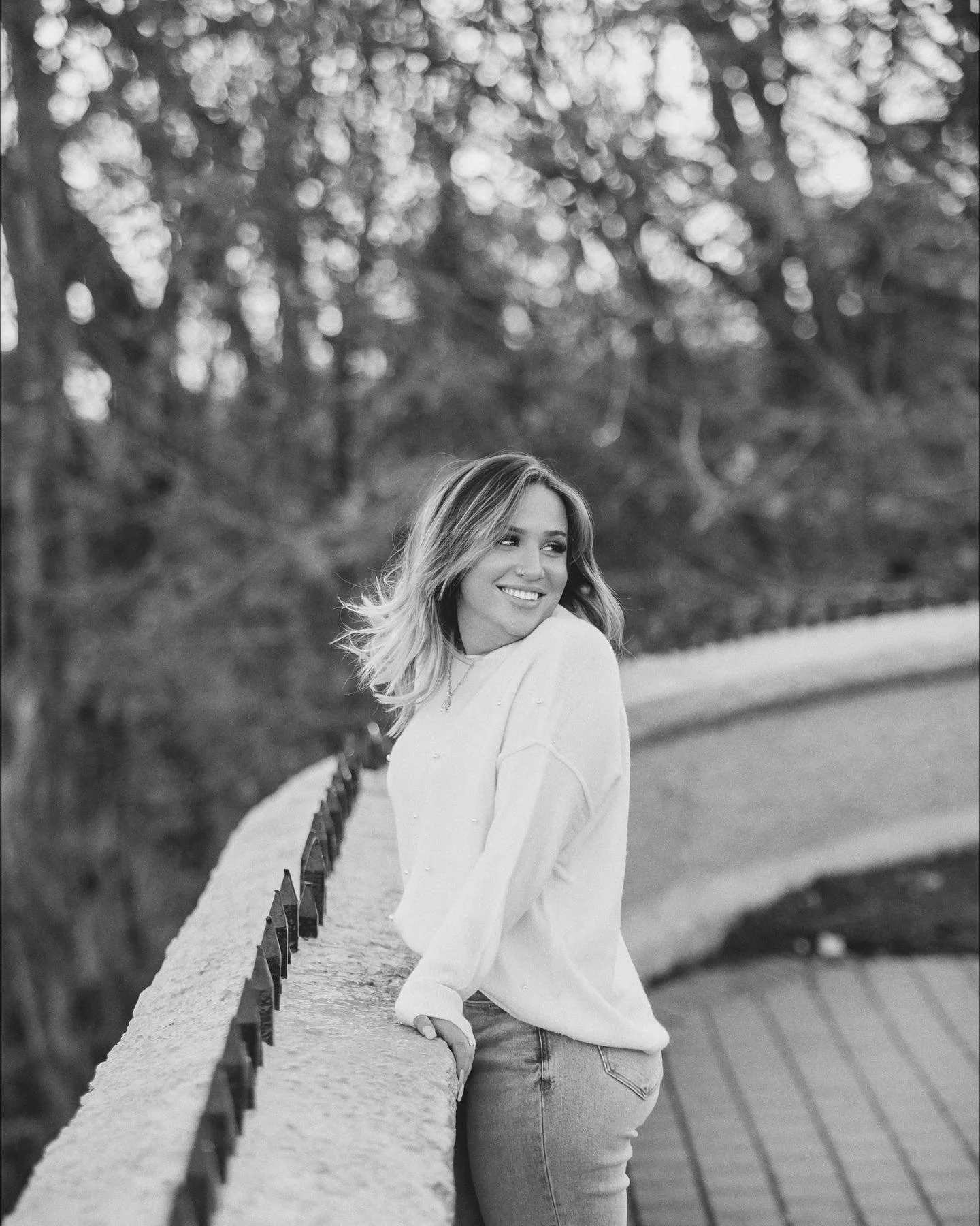 A young woman with wavy hair standing outdoors, leaning on a curved stone railing, smiling and looking to the side, with trees in the background.