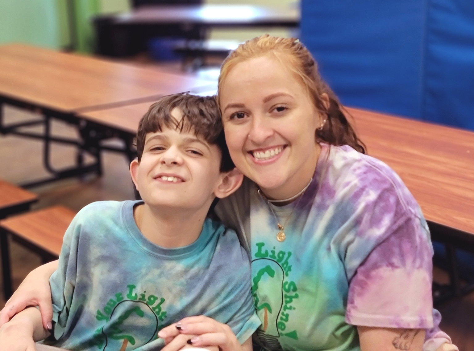 A young boy and a young woman smiling and sitting close together at a table in a room, wearing tie-dye shirts with green text and a graphic, with tables and chairs in the background.