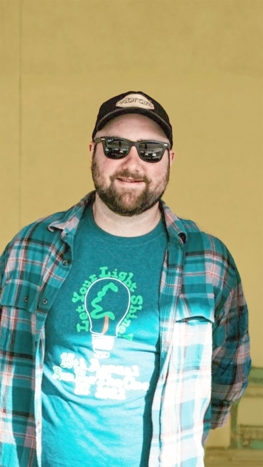 A behavioral therapist man with a beard and sunglasses smiling, wearing a plaid shirt over a teal t-shirt with a lightbulb graphic that says "Let Your Light Shine 15th Annual Run for the One 2022."