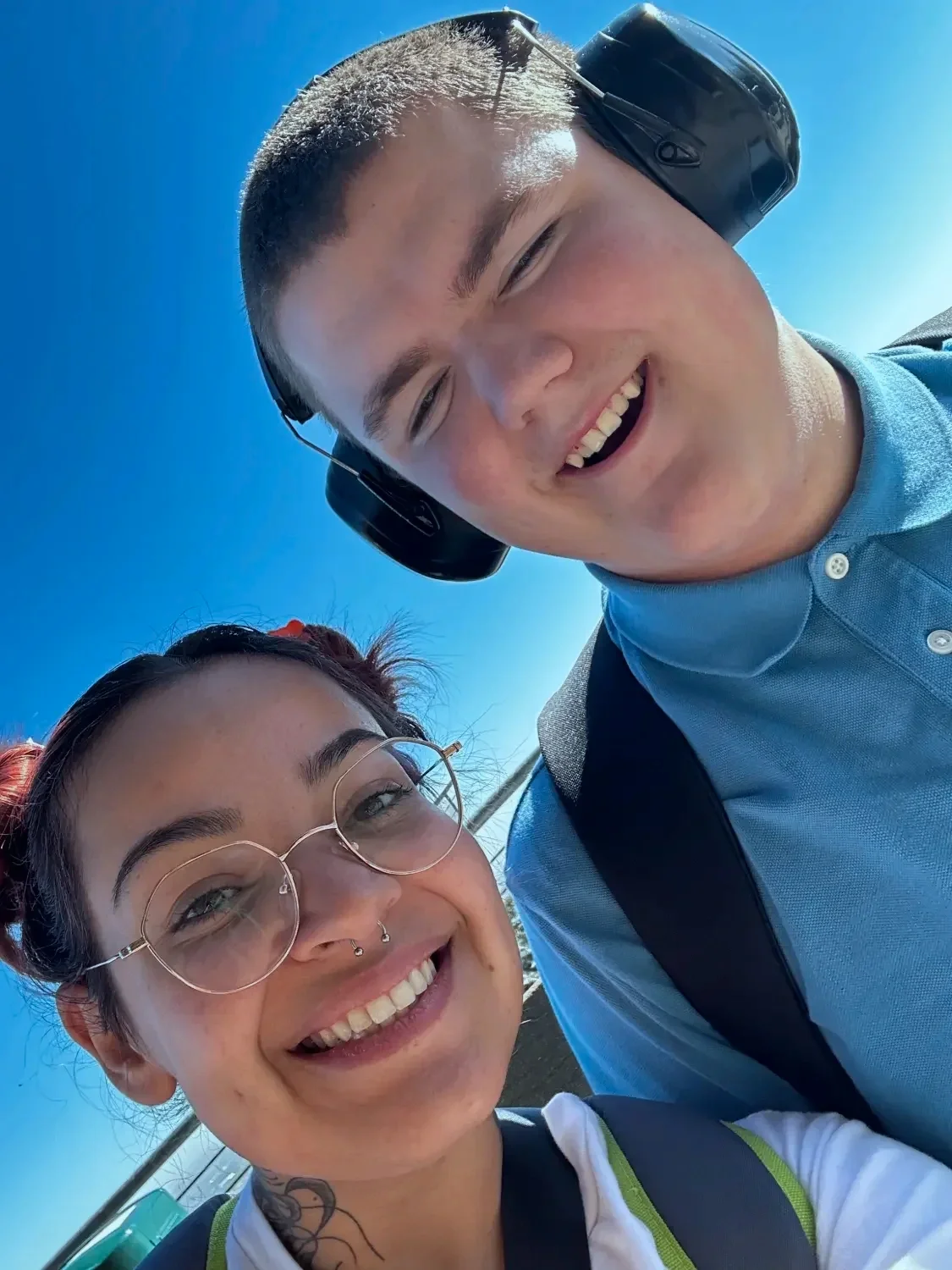 A young man and woman are smiling for a selfie outdoors against a clear blue sky. The man is wearing a blue polo shirt, black headphones, and carries a backpack. The woman has glasses, a septum piercing, and red hair with a tattoo on her neck.