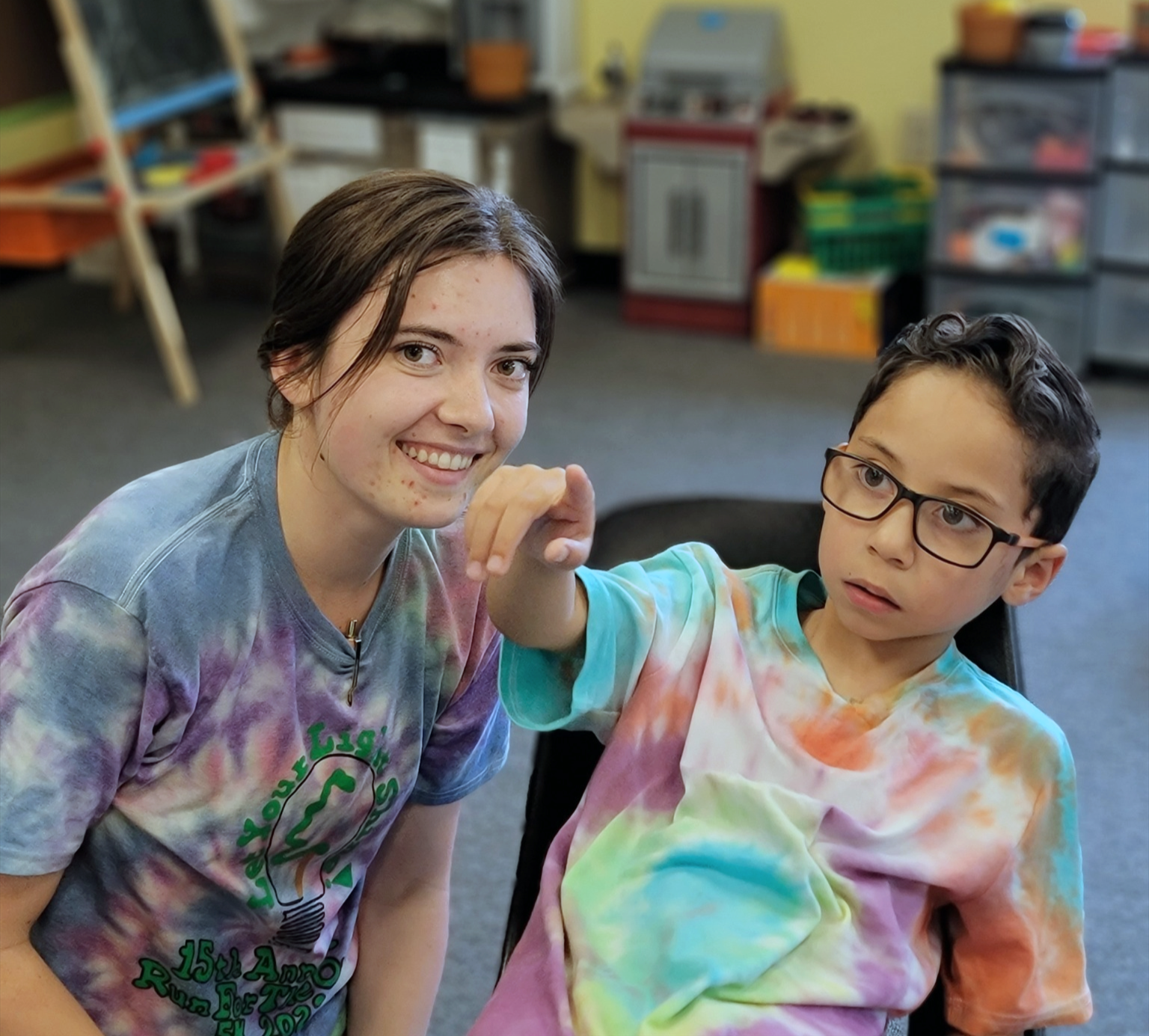 A behavioral therapist woman and a boy with glasses sitting together. The girl is smiling at the camera, and the boy is pointing off-camera. Both are wearing tie-dye shirts with a lightbulb logo.