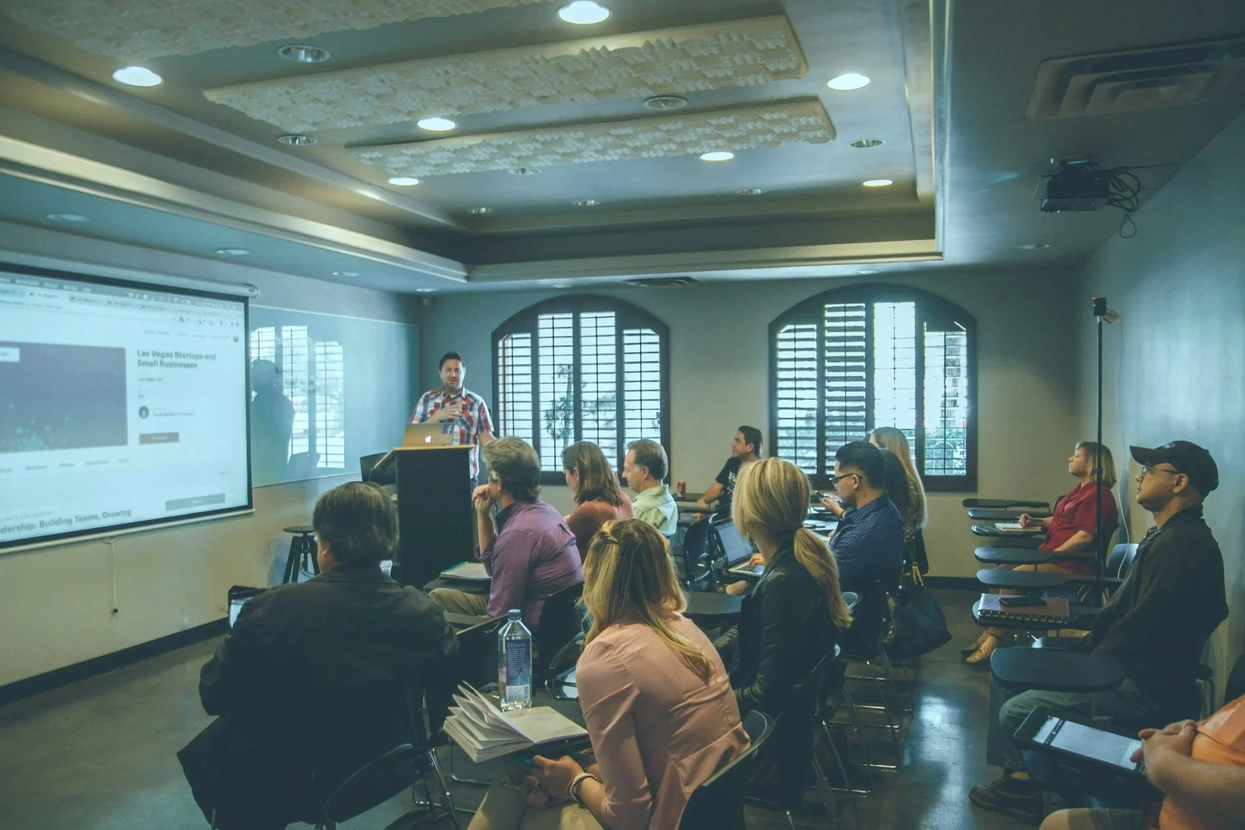 A man giving a presentation to an audience in a conference room with large windows and natural lighting.