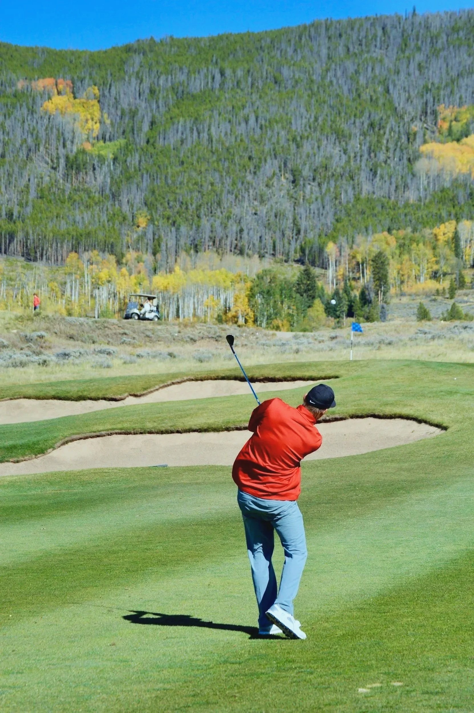 A golfer in a red shirt swinging a club on a mountain course, with bunkers, pine trees, and fall foliage visible in the background.
