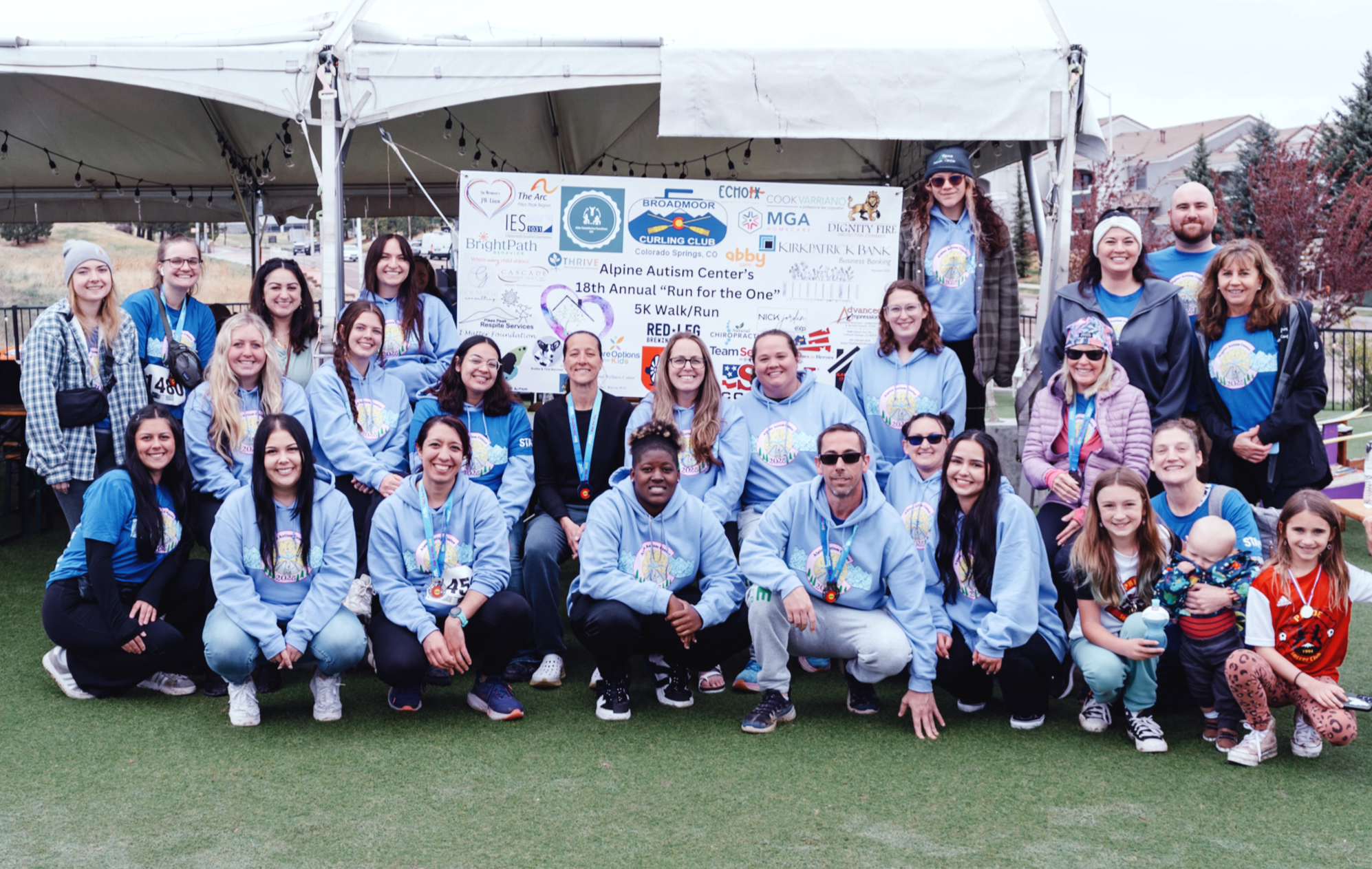 large group of about 30 adults and a few children posing for a group photo outdoors in front of a white tent and a large sign for the "Alpine Autism Center's 18th Annual 'Run for the One' 5K Walk/Run." Most adults are wearing light blue hoodies.