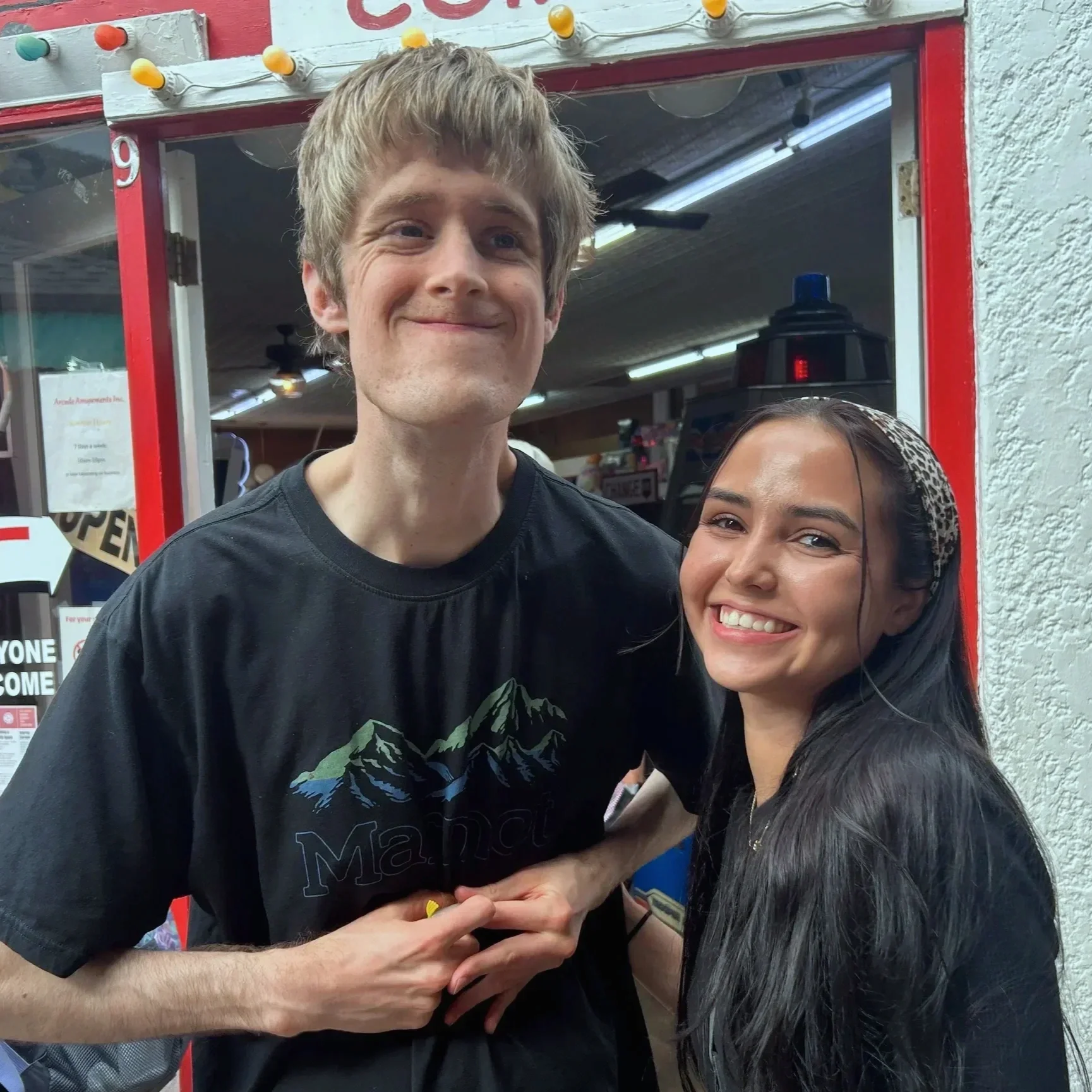 A man and woman smiling and posing together inside a store with a red and white storefront in the background.