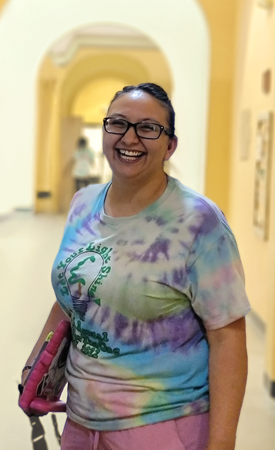 A smiling behavioral therapist woman wearing glasses and a tie-dye shirt with a lightbulb graphic that says "Let Your Light Shine," standing in a hallway.
