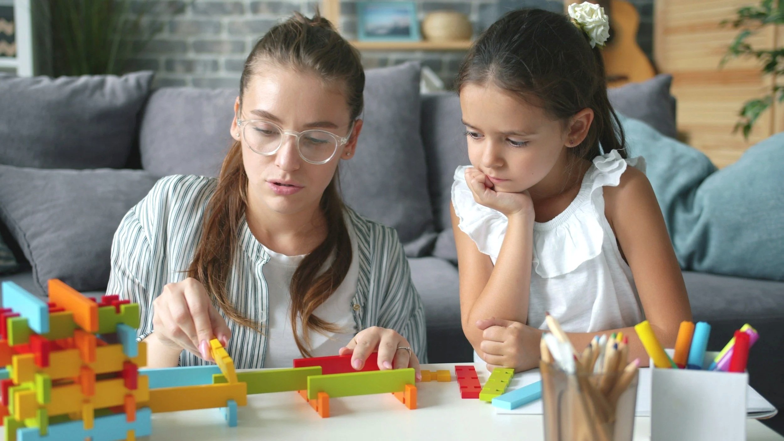 A woman with glasses and a young girl looking intently at colorful, interlocking building blocks on a table.