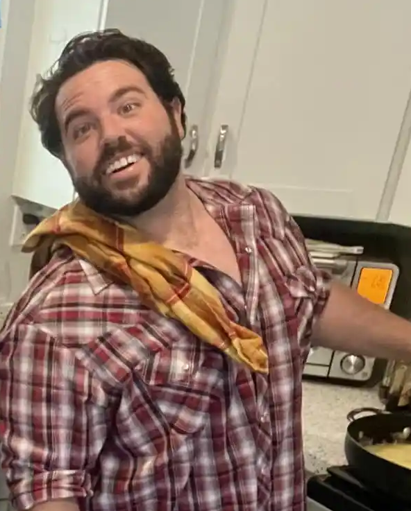 A man with dark curly hair and a beard wearing a red plaid shirt and a yellow bandana around his neck, smiling while cooking in a kitchen.