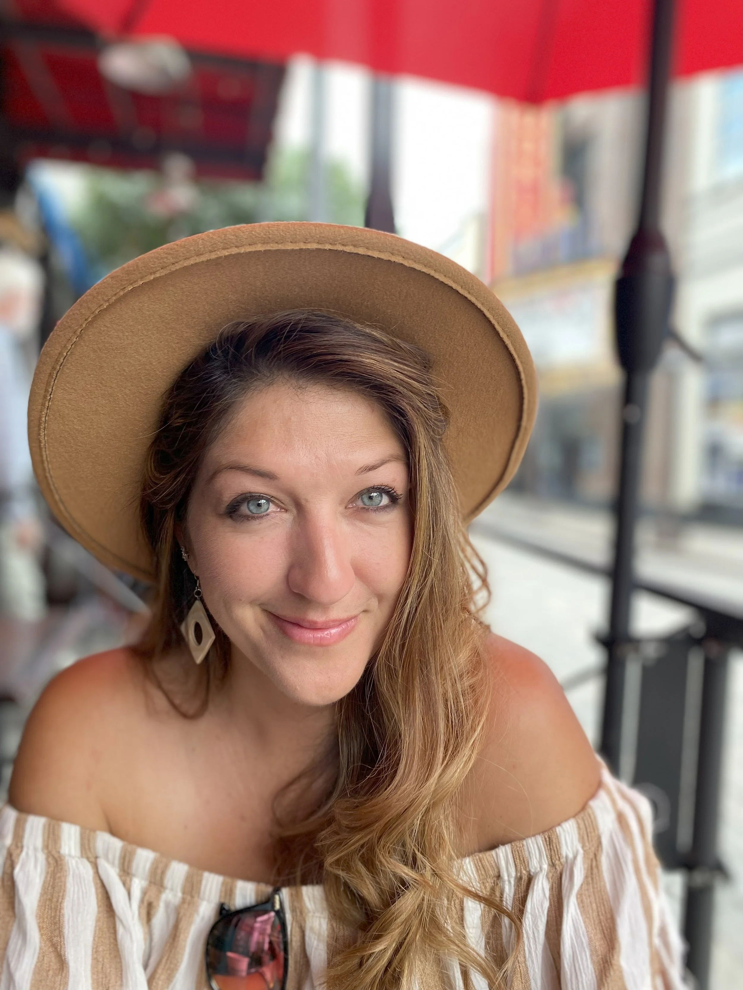 A woman with blue eyes, wearing a wide-brimmed tan hat and earrings, smiling at the camera, with long wavy hair, in an outdoor setting with a red canopy above and city buildings in the background.