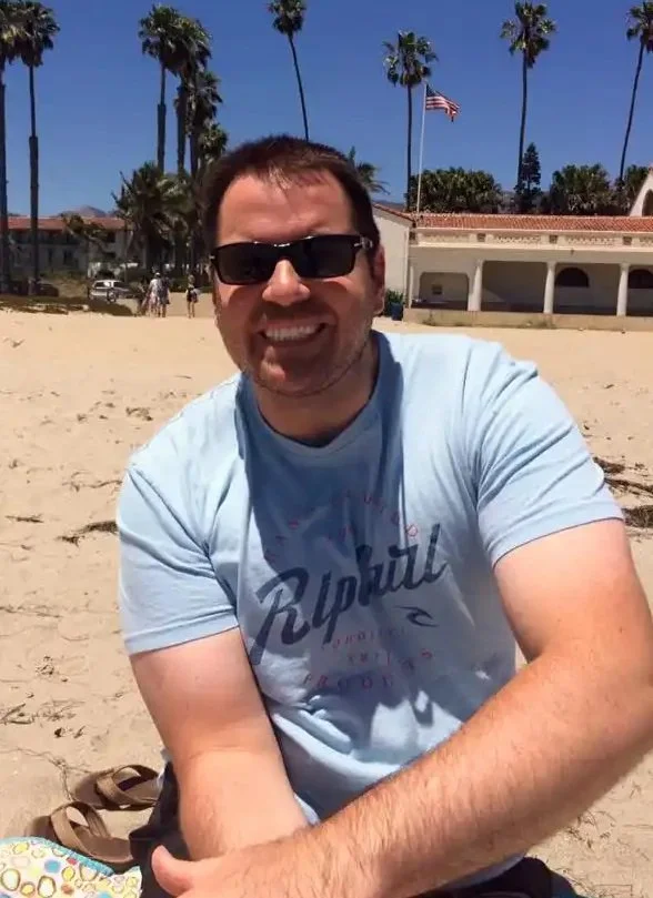 Smiling man wearing sunglasses and a light blue T-shirt with text, sitting on sandy beach with palm trees, American flag, and building in background.