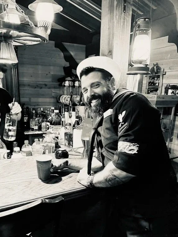A bearded man smiling and leaning on a bar with liquor bottles in the background, wearing a chef's hat and dark apron, in a rustic wooden setting.