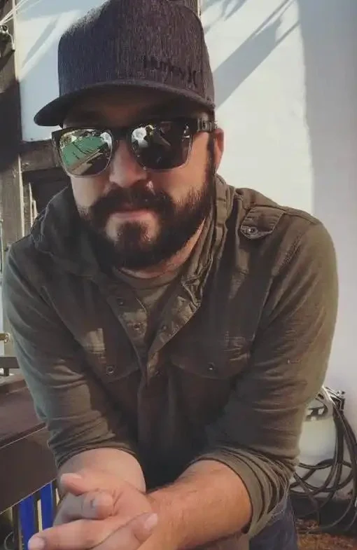 A man with a beard and sunglasses wearing a black cap and a button-up shirt, leaning forward with his arms crossed on a table or counter.