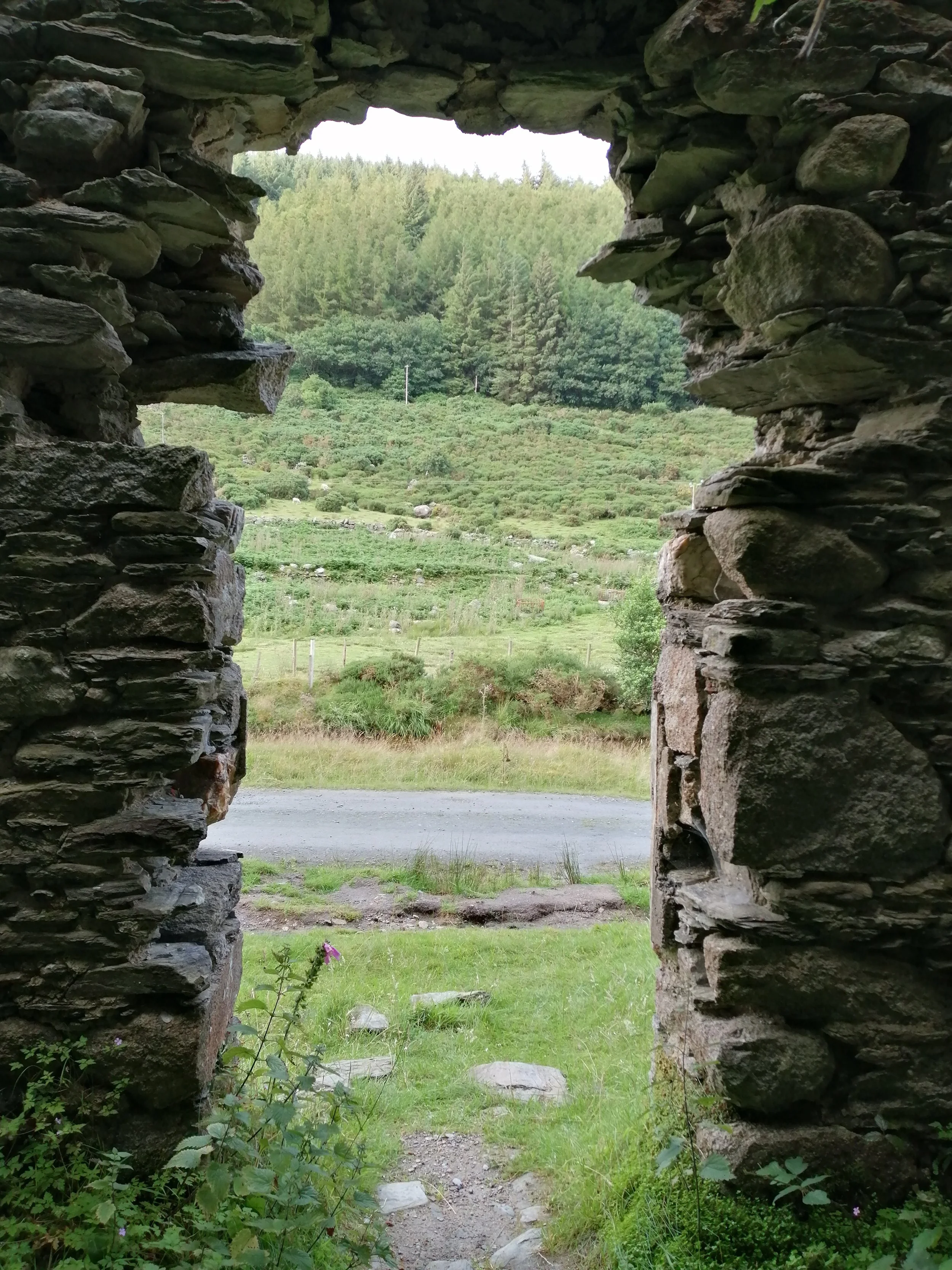 Looking through a rough stone doorway towards grass, a narrow road and a wooded hillside.