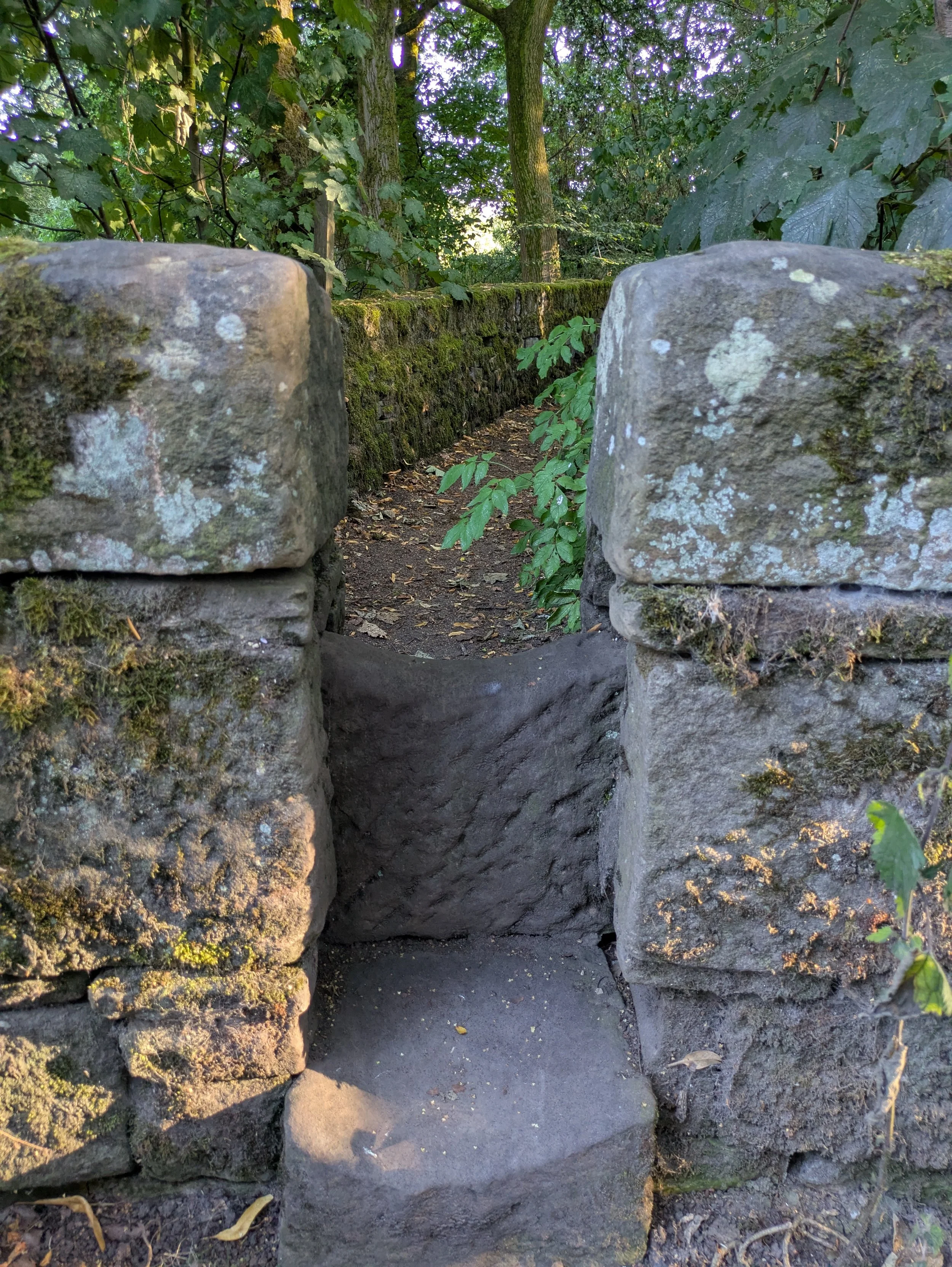 A narrow stone passage leading onto a shaded woodland path between moss-covered walls.