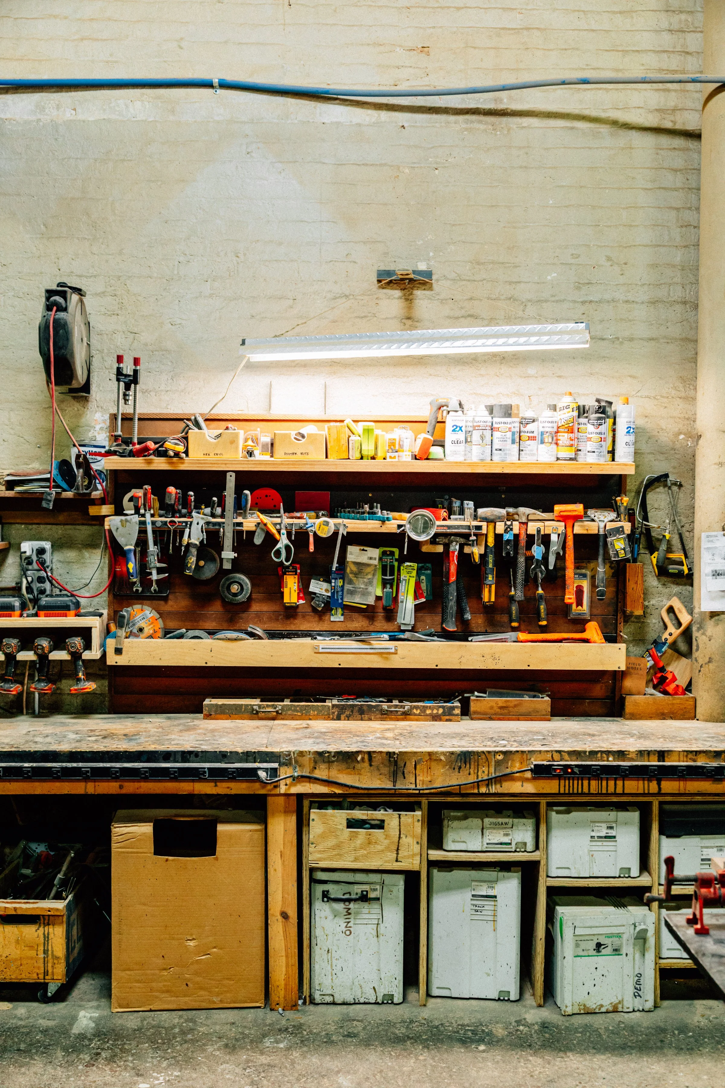 A workbench in a workshop with various tools, paint cans, and storage containers, against a beige brick wall.