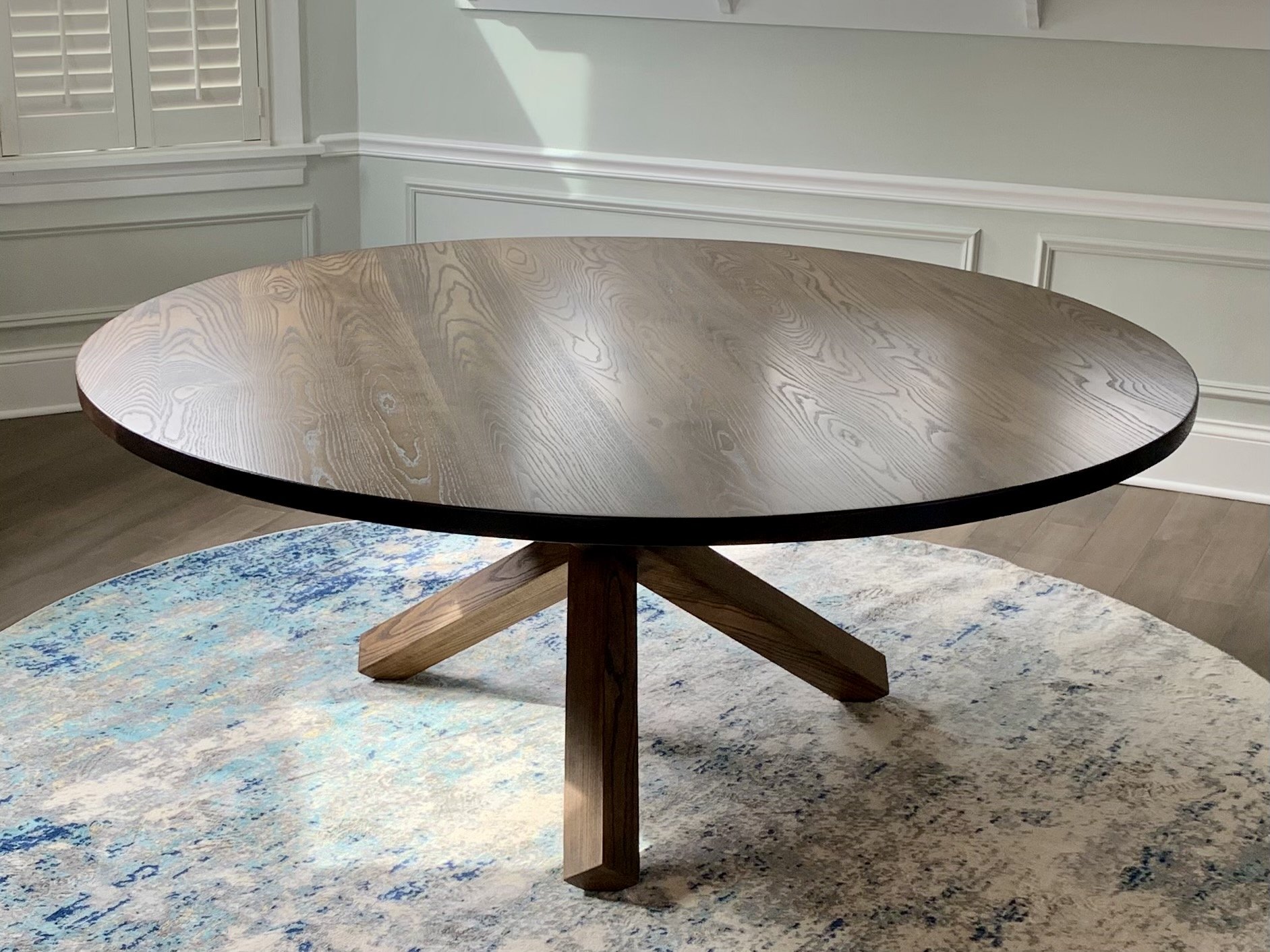 A round wooden dining table with a polished surface and a pedestal base, situated in a room with a patterned area rug and wainscoting on the walls.