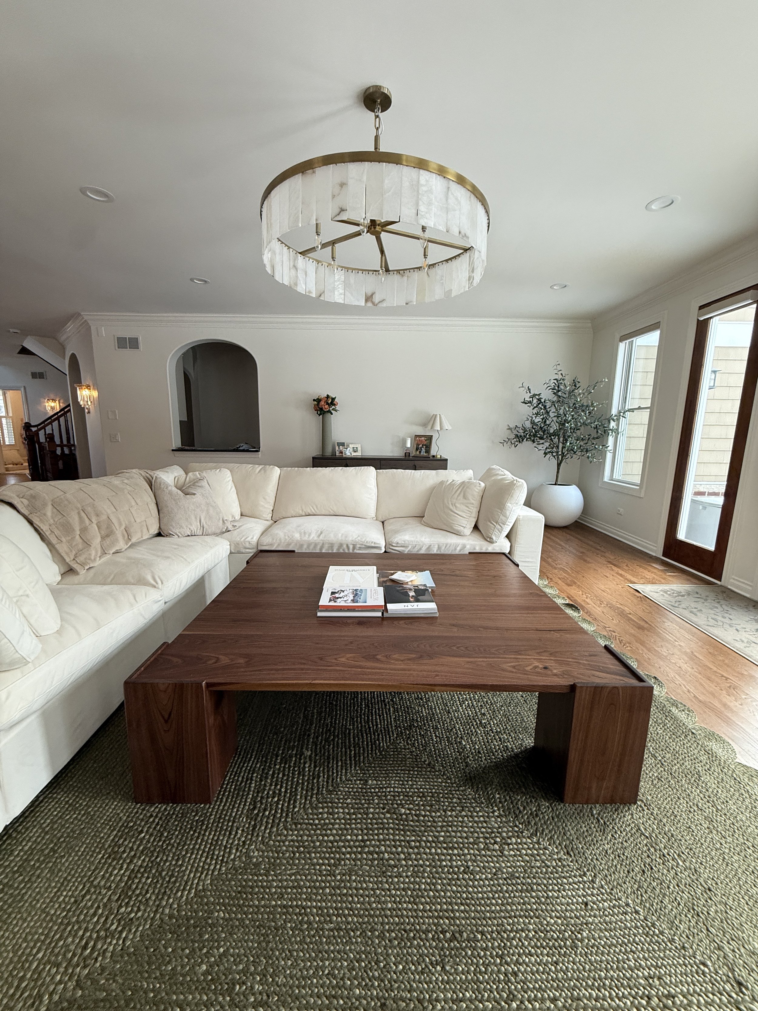 Living room with a round chandelier, beige sectional sofa, wooden coffee table, potted plant, framed photographs, and large windows.