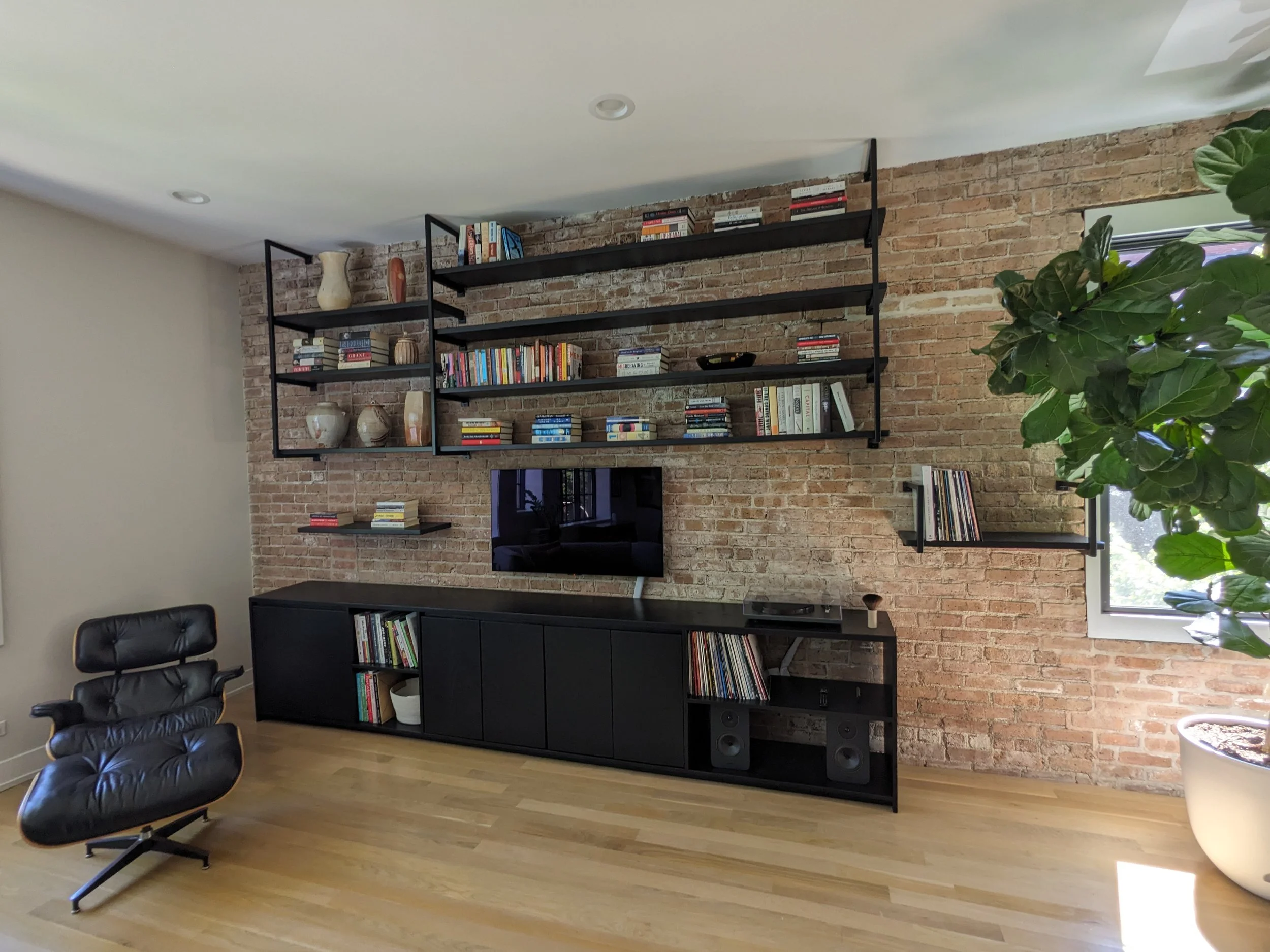 Interior view of a room with a brick accent wall, black shelving filled with books and decorative items, a flat-screen TV, a large green plant, a black office chair, and hardwood flooring.
