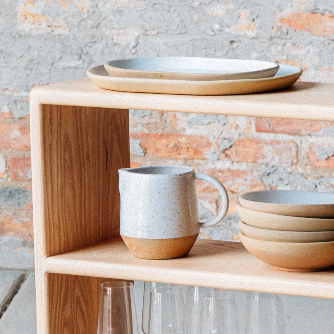 A wooden shelf holding ceramic plates, bowls, a mug, and glassware against a brick wall.