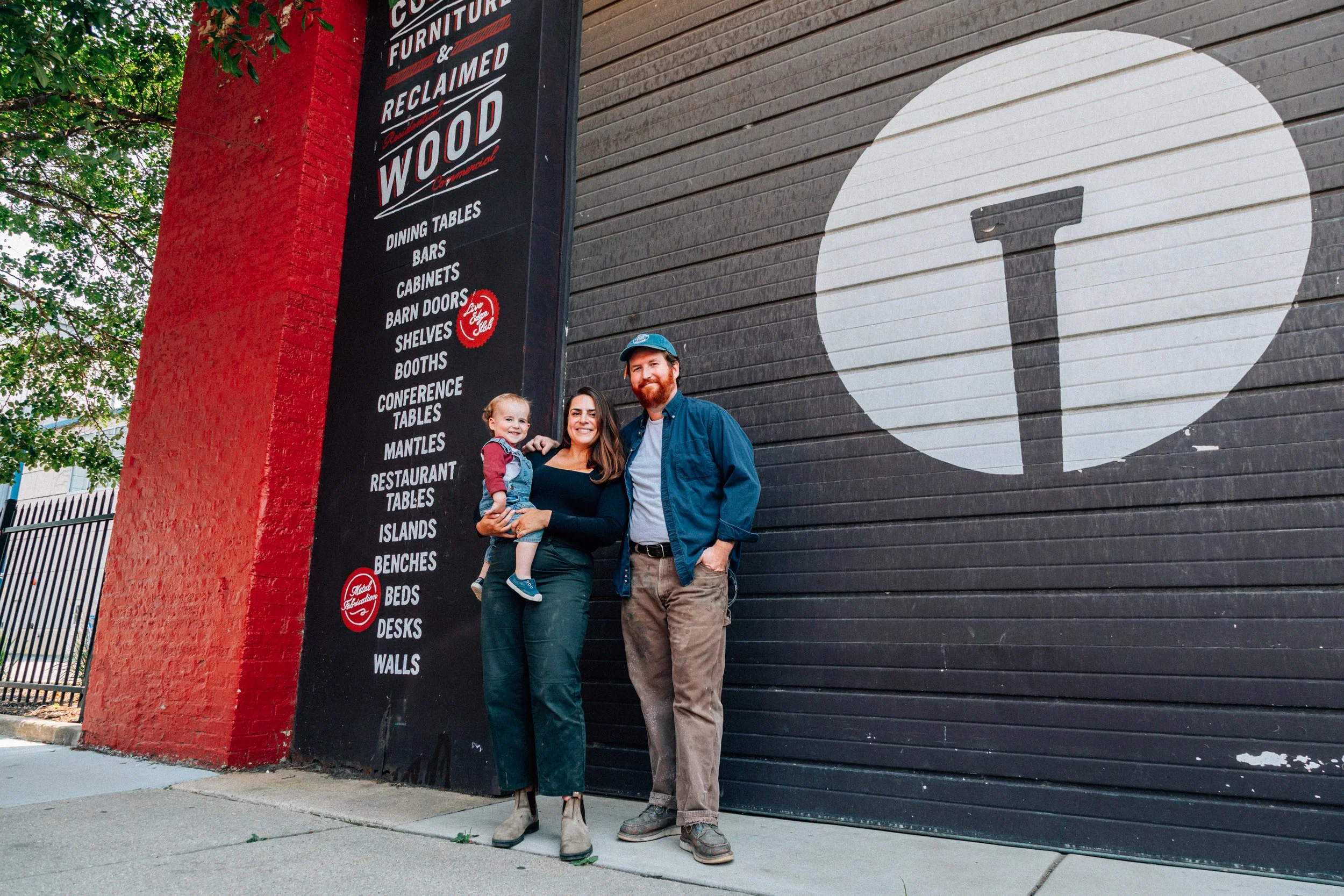 A family of three standing in front of a black and red wall with signage for furniture, including a large white letter 'T' in a circle. The woman is holding a young boy, both smiling, while the man stands next to them with his hand in his pocket.