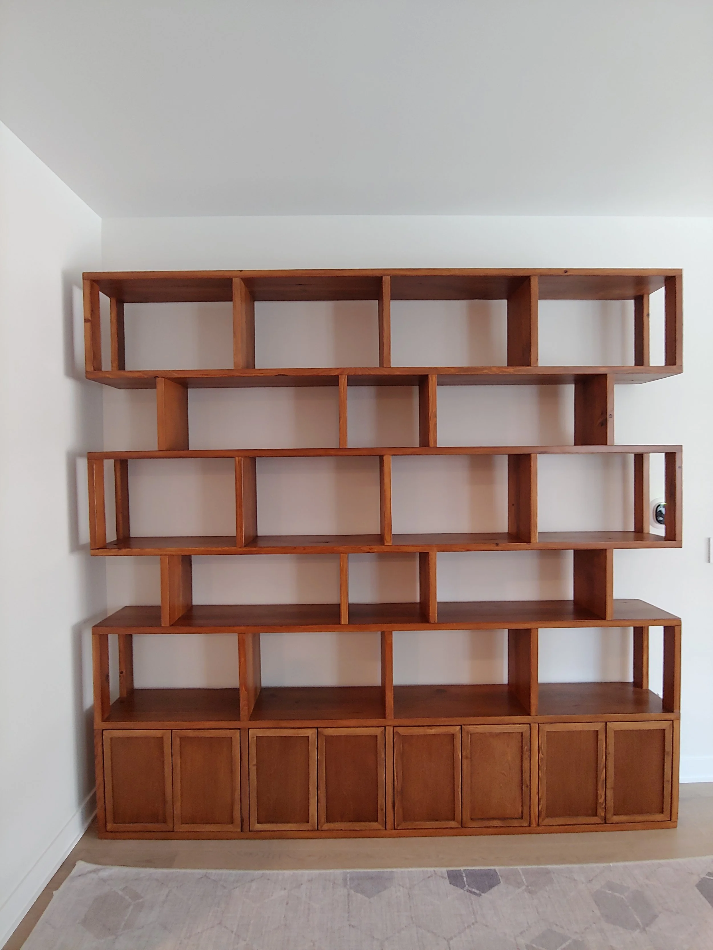 A large wooden shelving unit with multiple open compartments and closed cabinets at the bottom, mounted on a white wall in a room with light-colored flooring and a patterned rug.