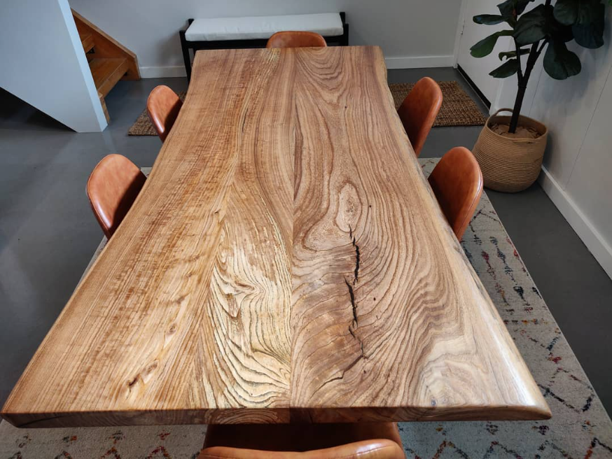 A large wooden dining table with a natural, live edge finish surrounded by five brown leather chairs. In the background, there is a small bench, a potted plant, and a patterned rug.