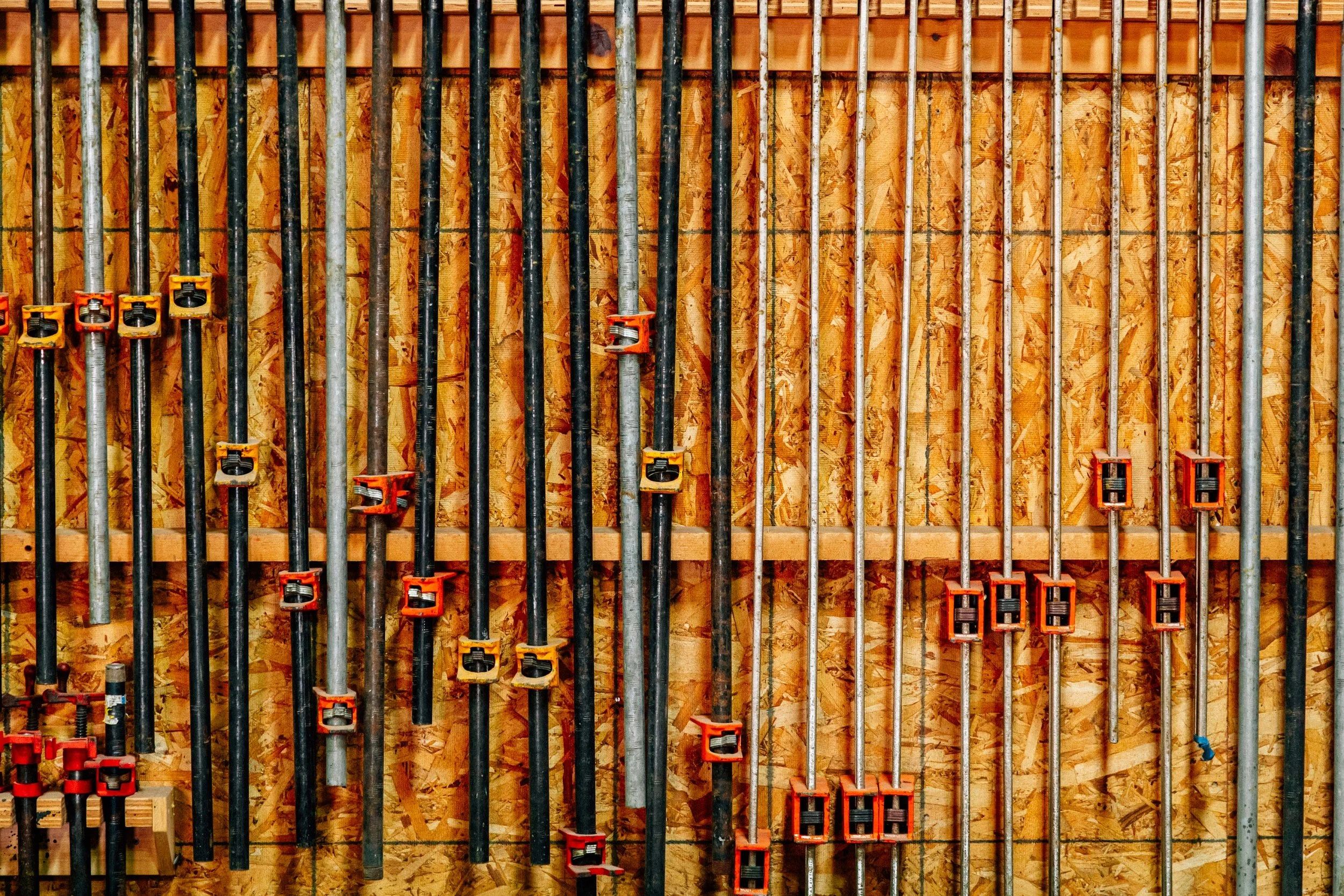 Array of metal pipes with orange clamps mounted on a wooden wall in a construction setting.